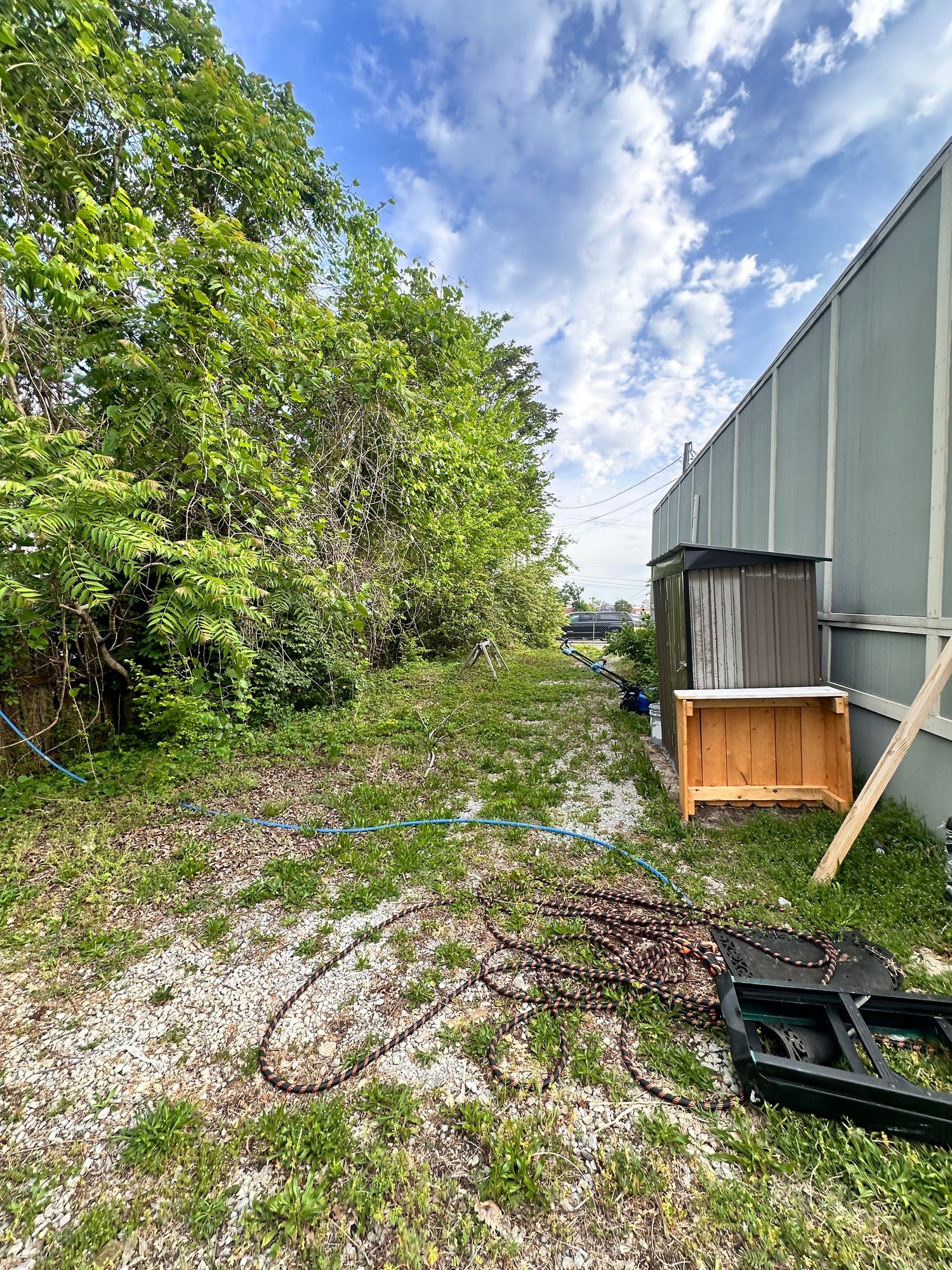 Overgrown vacant lot beside a metal building with scattered chains, a wooden crate, and lush green trees under a cloudy sky.