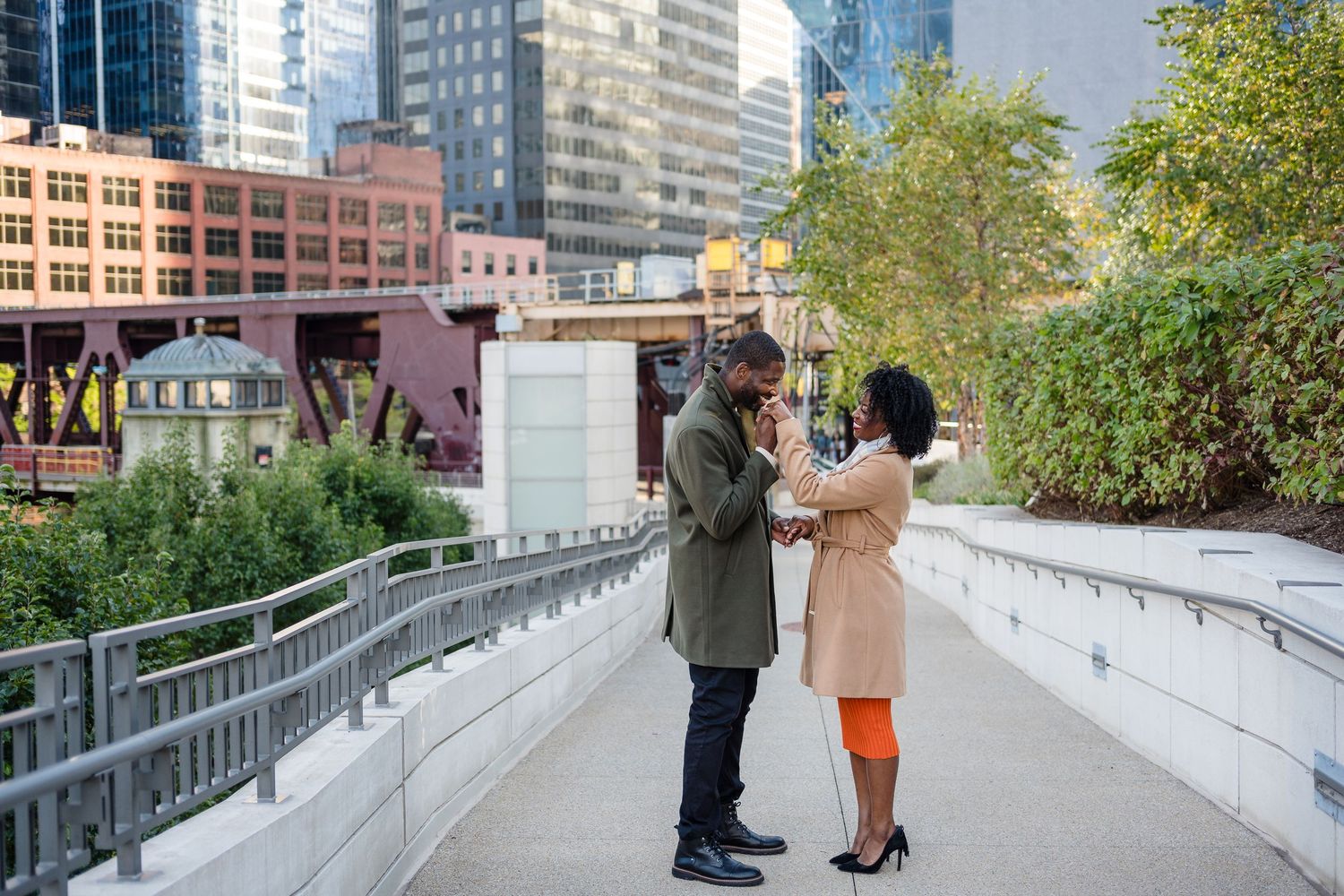 A couple shares a romantic moment on a city walkway surrounded by urban architecture and lush greenery.