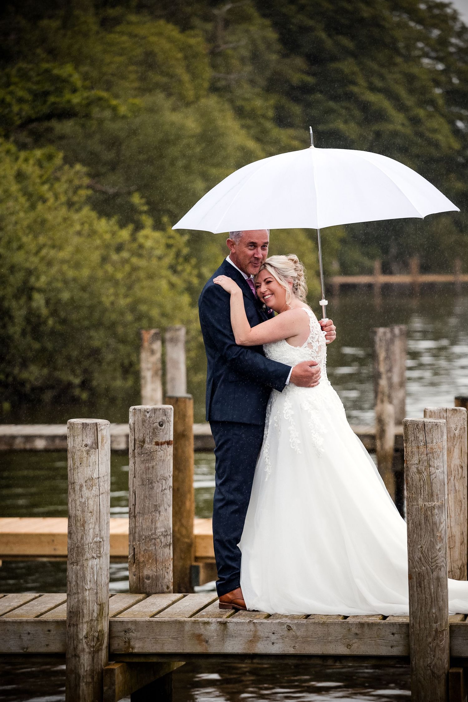 A bride and groom embrace under a white umbrella on a wooden dock by a lake on their rainy wedding day.