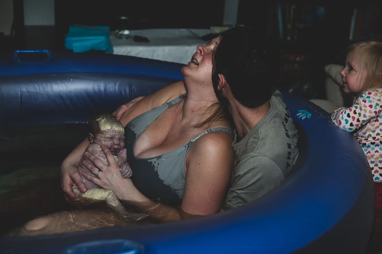 A supported labor scene in a blue inflatable birthing pool at home.