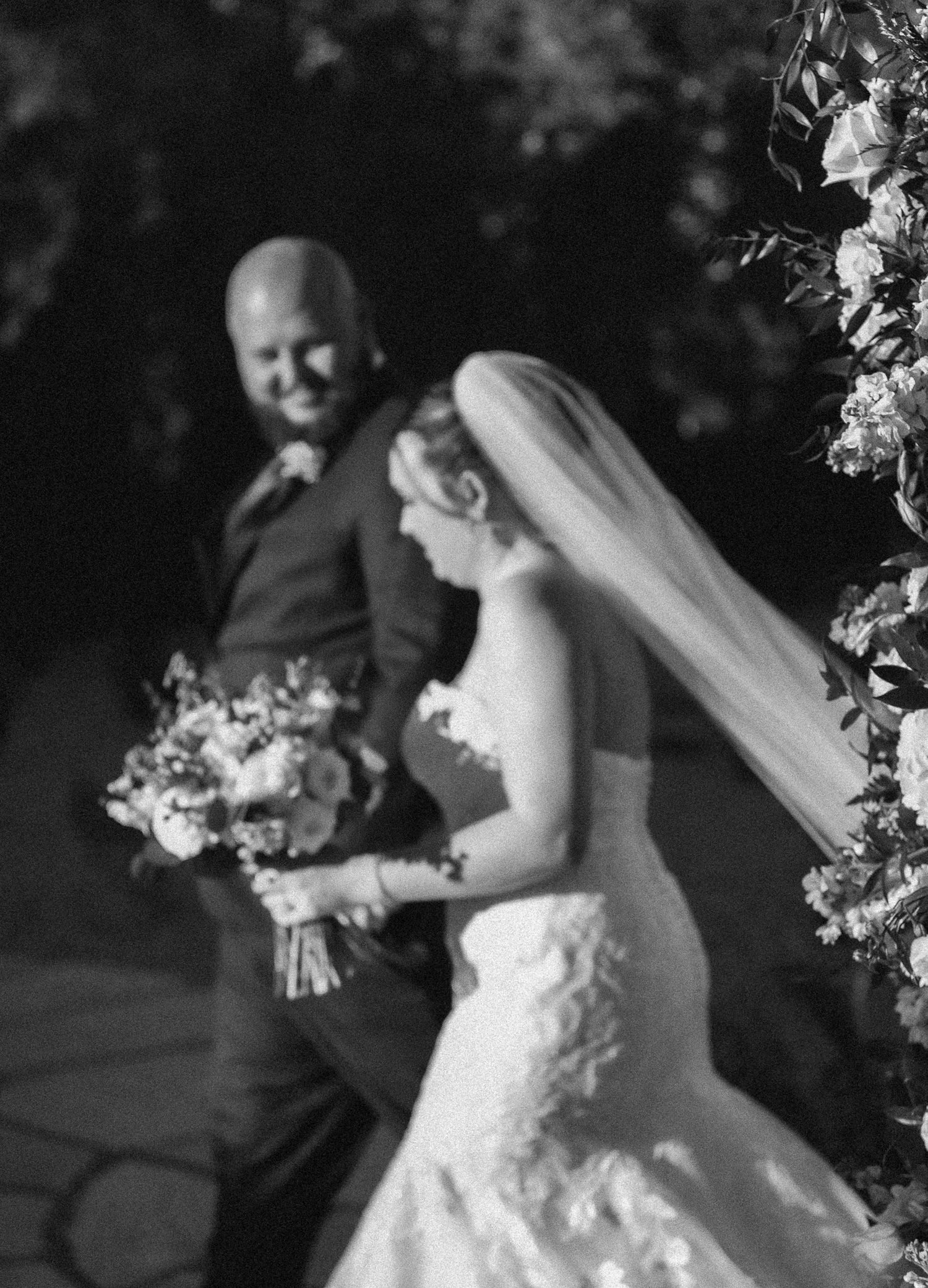 Black and white photo of a bride holding a bouquet walking with a bald groom near a floral arch at a wedding ceremony.