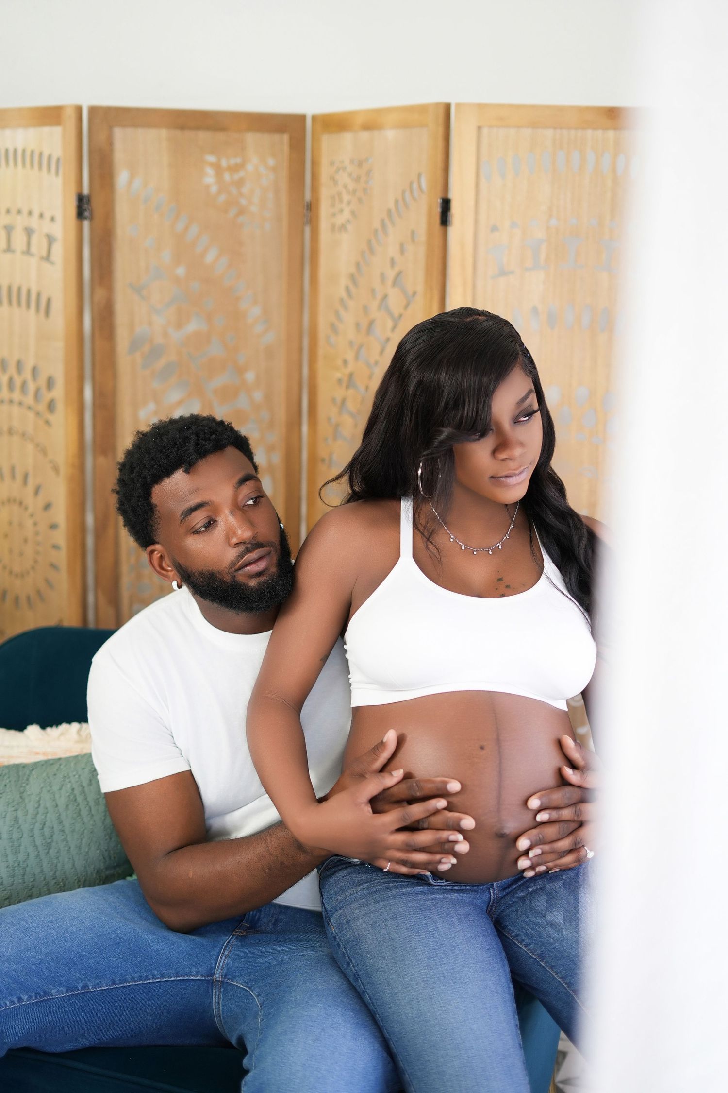 Pregnant woman and her husband  sit together on a blue couch with a decorative folding screen in the background.