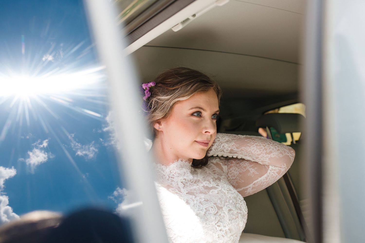 Bride in lace wedding dress adjusting her hair inside a car, with sunlight and blue sky visible through the window at Bull Point Estate