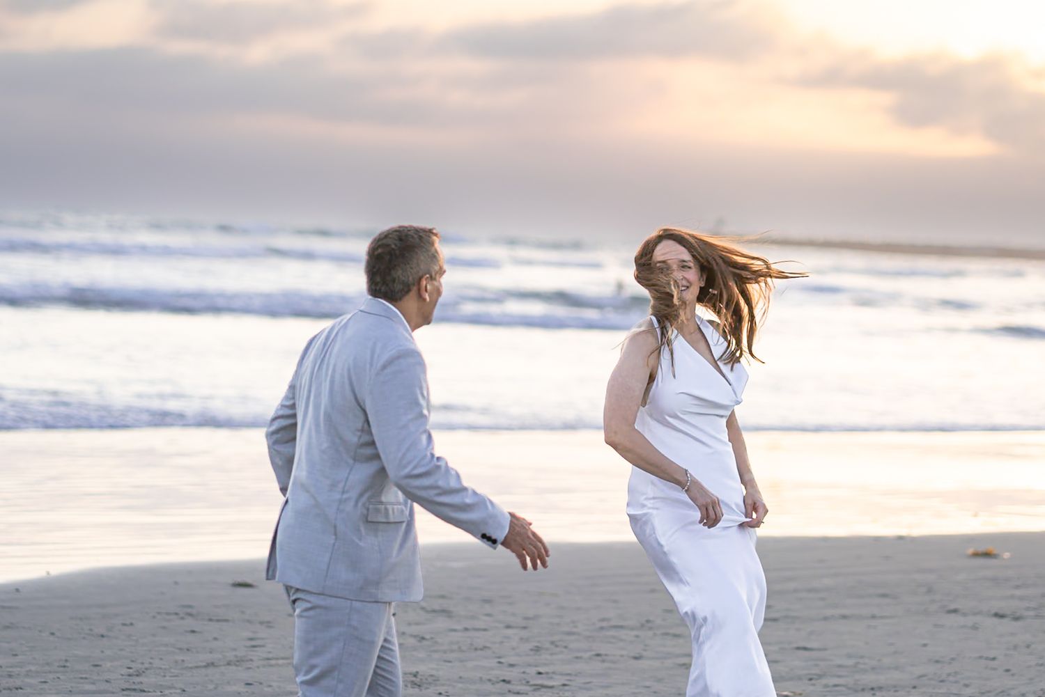 Couple dancing joyfully on a sandy beach at sunset, woman in white dress with flowing red hair, man in light gray suit. Oceanside, CA