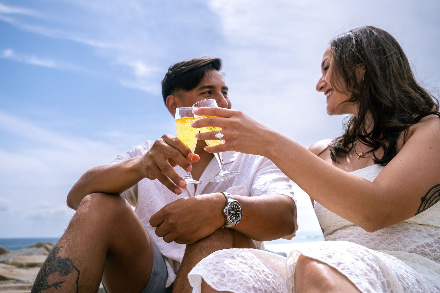 Newly engaged couple toasting with champagne glasses on a sunny beach, smiling and displaying the ring. Carlsbad, CA