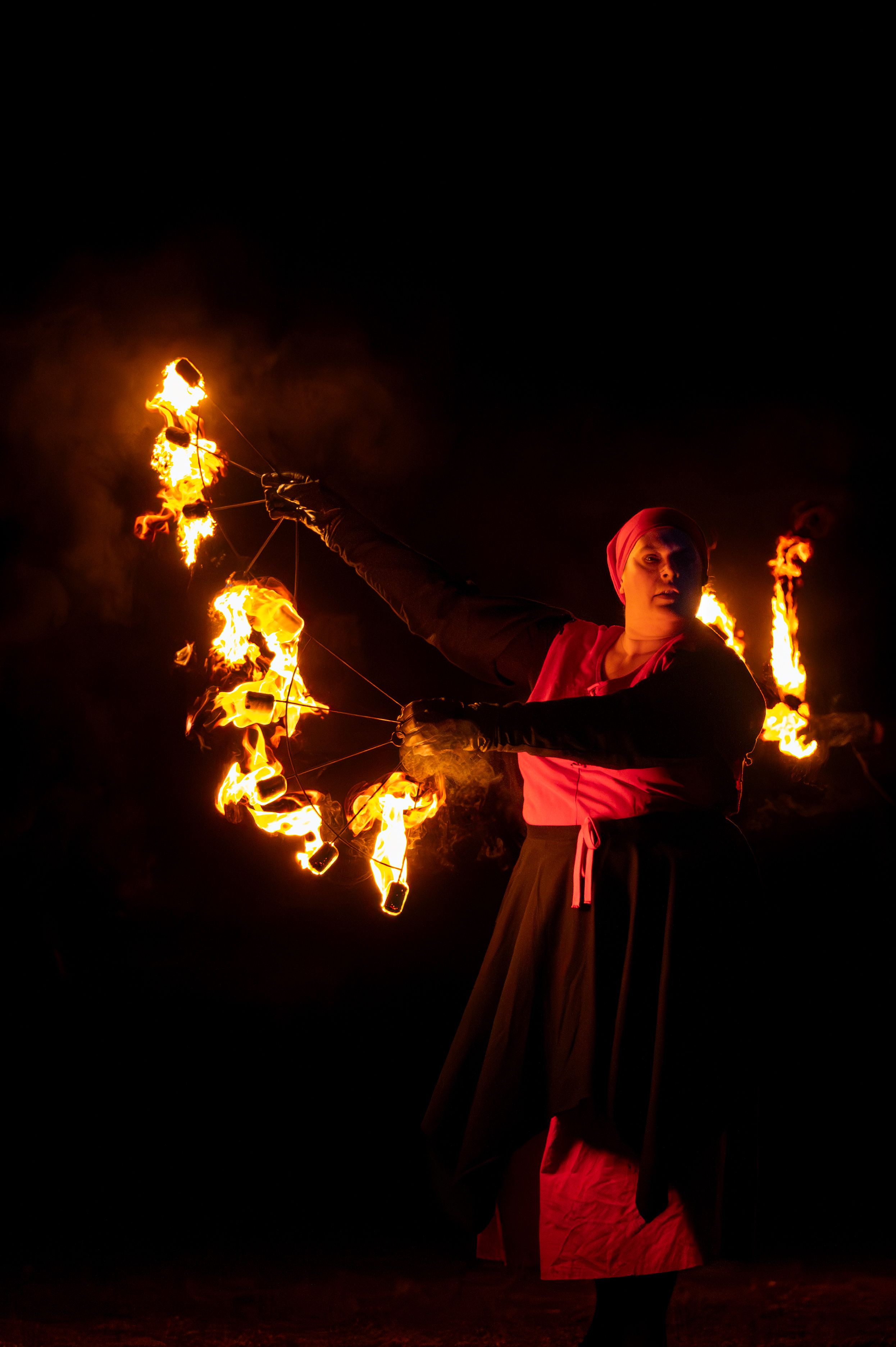 Un artiste du feu faisant tournoyer des éventails de feu lors d'un spectacle nocturne en plein air, dans une atmosphère enfumée.