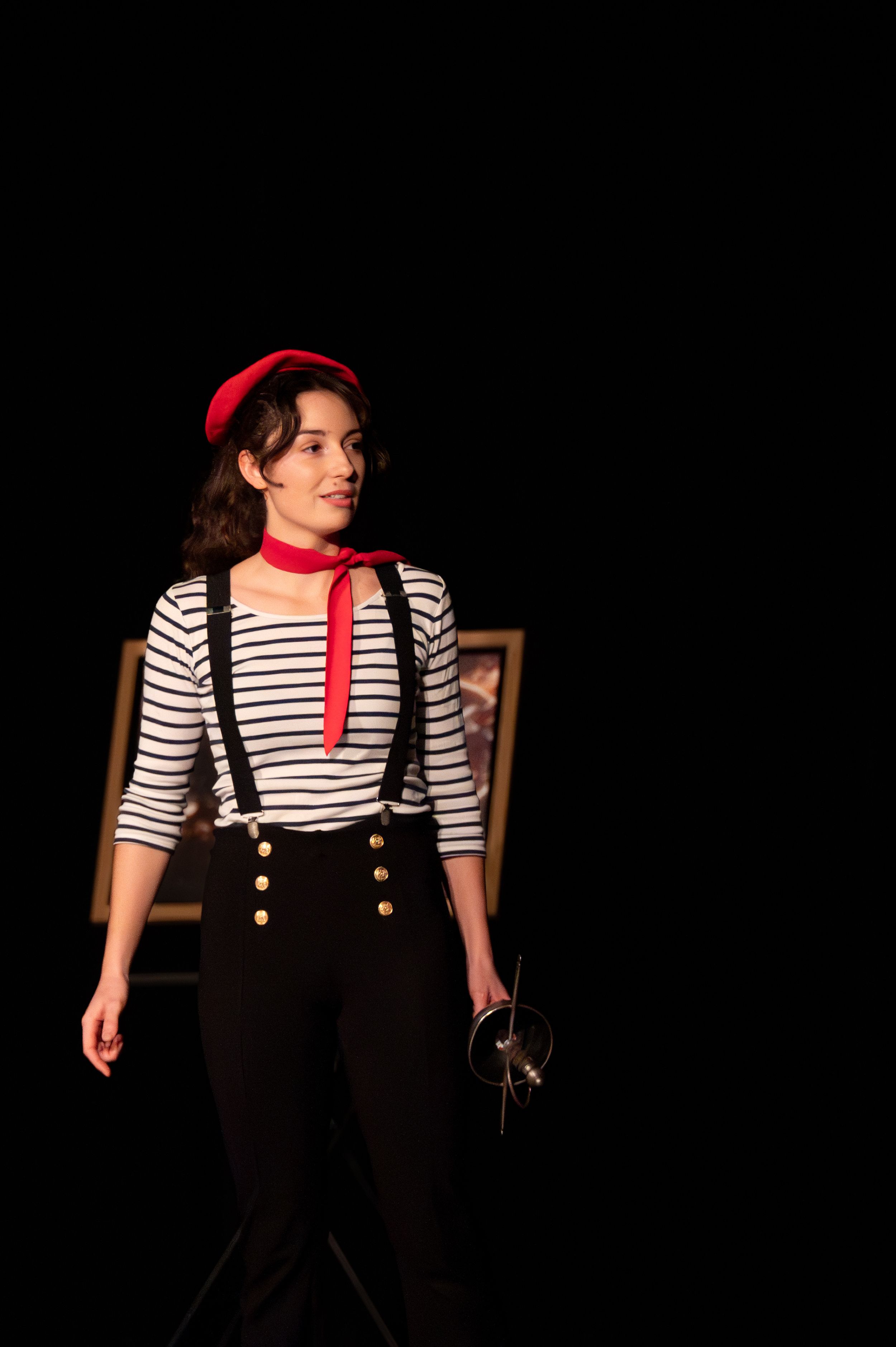 Une jeune femme vêtue d'un costume d'inspiration française, avec un béret rouge, un haut rayé, des bretelles et une écharpe rouge, sur scène