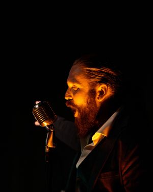 Un présentateur barbu se produisant devant un micro vintage, baigné d'une lumière dorée et chaleureuse sur un fond sombre.