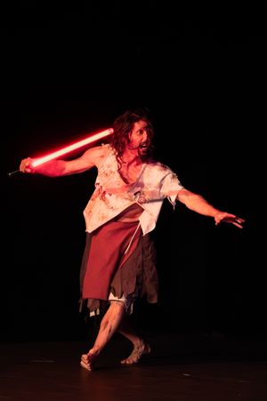 Un artiste vêtu d'un costume en lambeaux, brandissant un sabre laser rouge vif sur une scène sombre. Spectacle d'escrime artistique