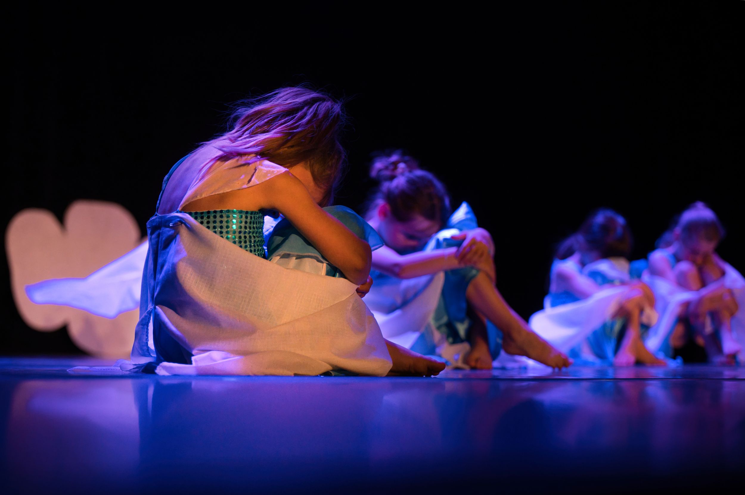 De jeunes danseuses vêtus de costumes blancs, assis sur une scène éclairée de bleu lors d'un spectacle de danse.