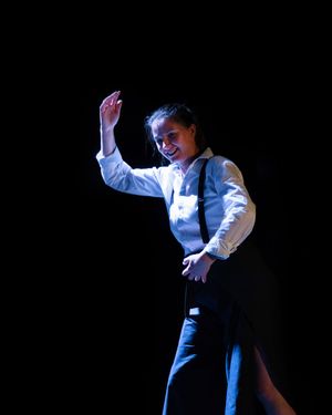 Une danseuse vêtue d'un gilet et de bretelles se produit avec le sourire sur une scène sombre.
