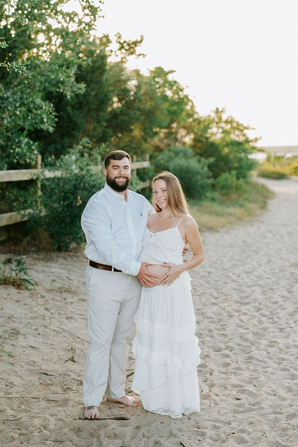 Pregnant couple in white outfits posing on sandy beach path surrounded by lush green trees during golden hour.