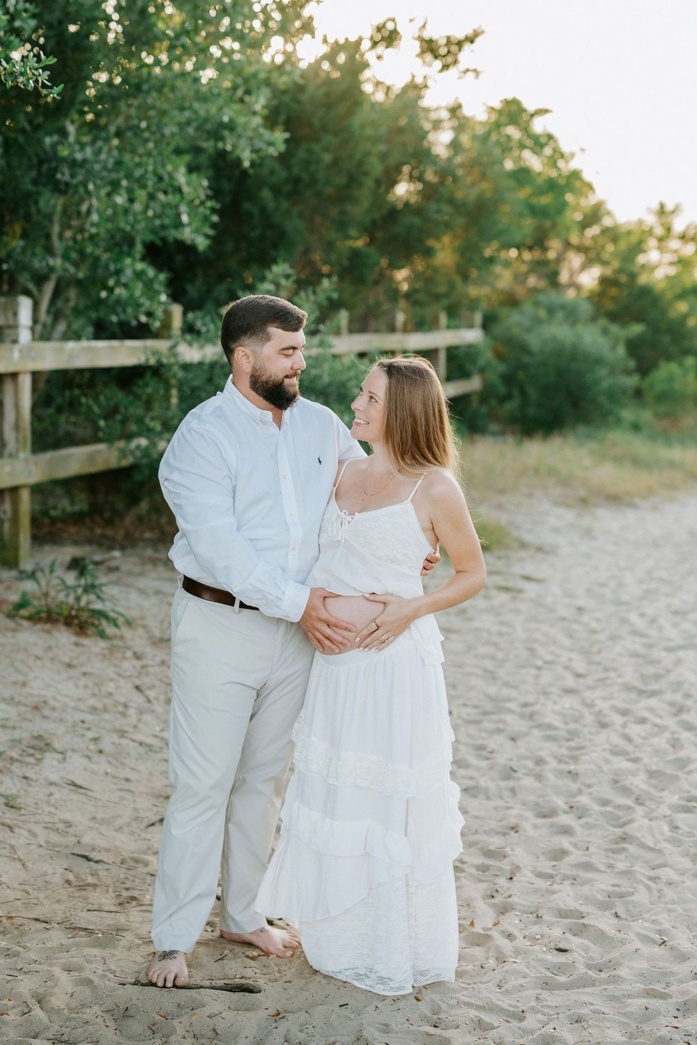 A couple in white outfits embraces on a sandy beach path, the woman visibly pregnant, surrounded by lush greenery.