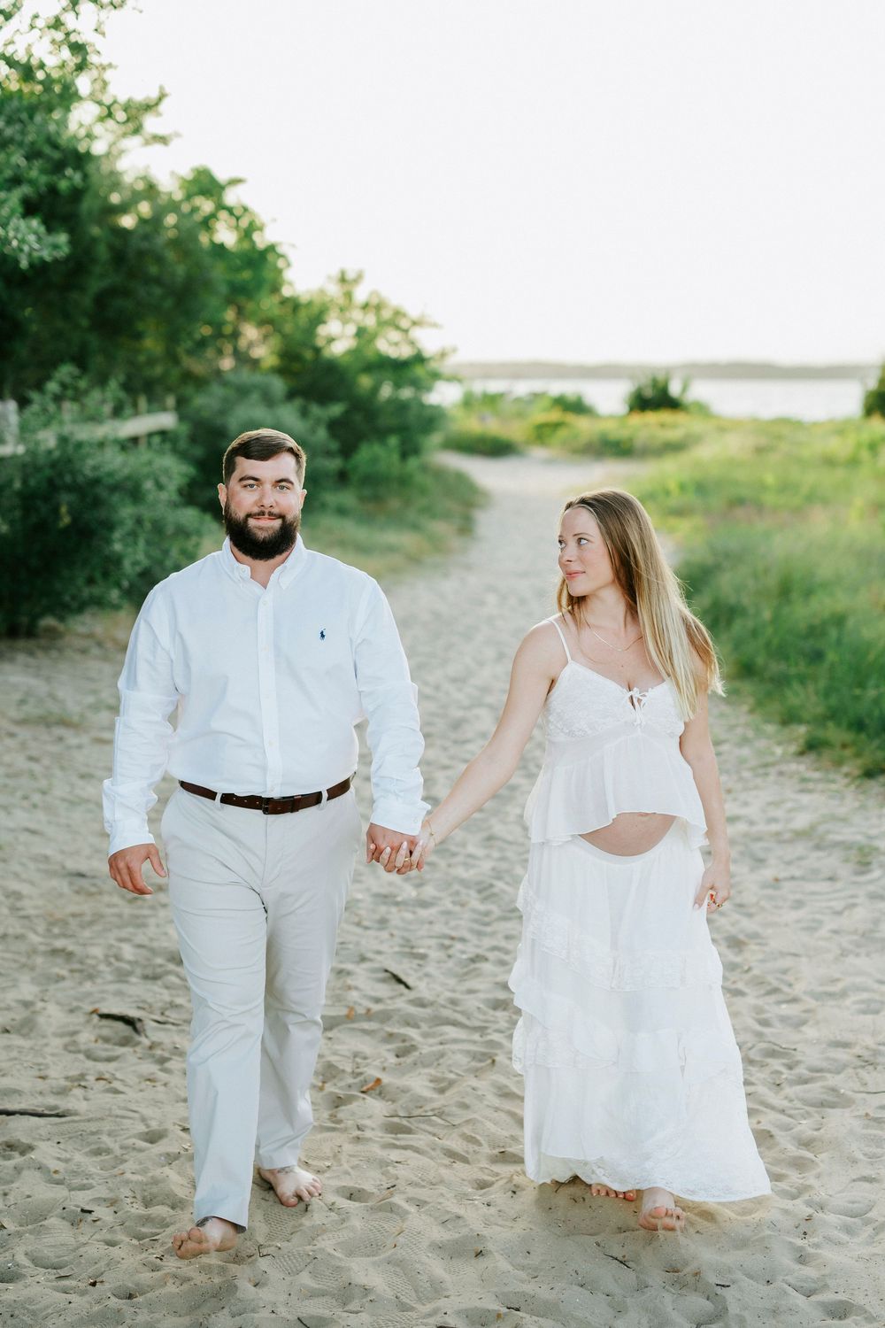 Pregnant couple walking hand-in-hand on sandy beach path, wearing white outfits, with green trees and water in background.