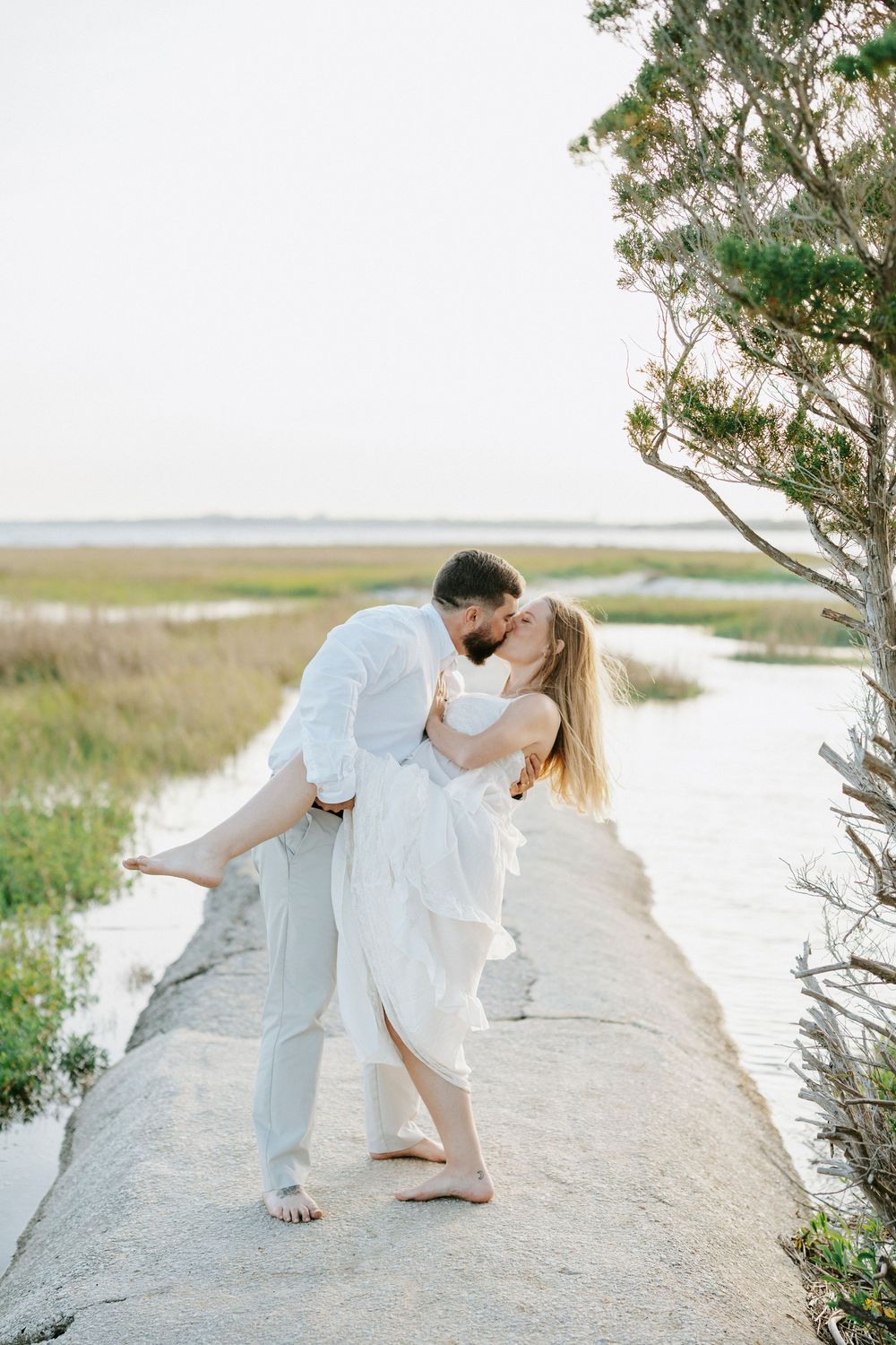 Couple sharing a romantic kiss on a coastal path, man dipping woman in white dress with marsh and water in background.