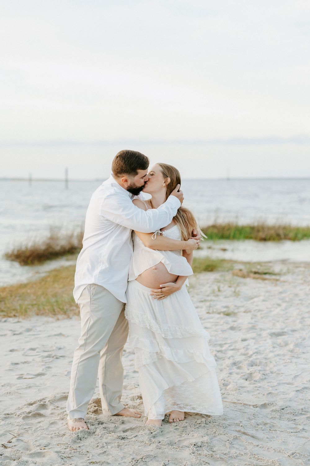 A couple shares a kiss on a sandy beach, with the woman in a white dress cradling her baby bump during a maternity photoshoot.