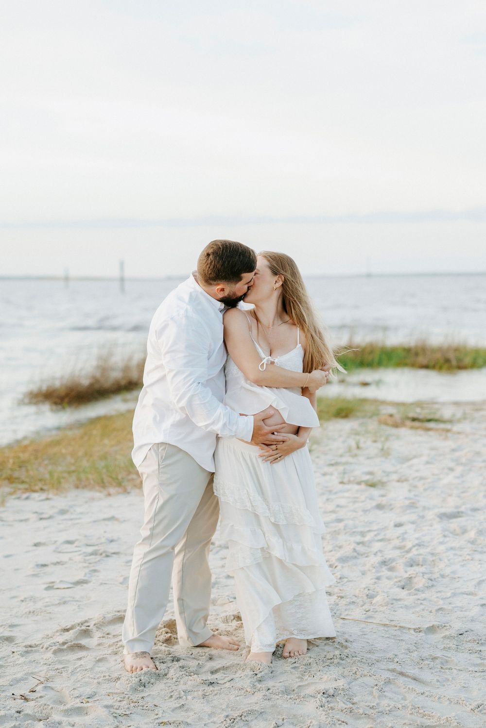 Couple sharing a romantic kiss on a sandy beach, woman in white maternity dress, man in white shirt and khaki pants.