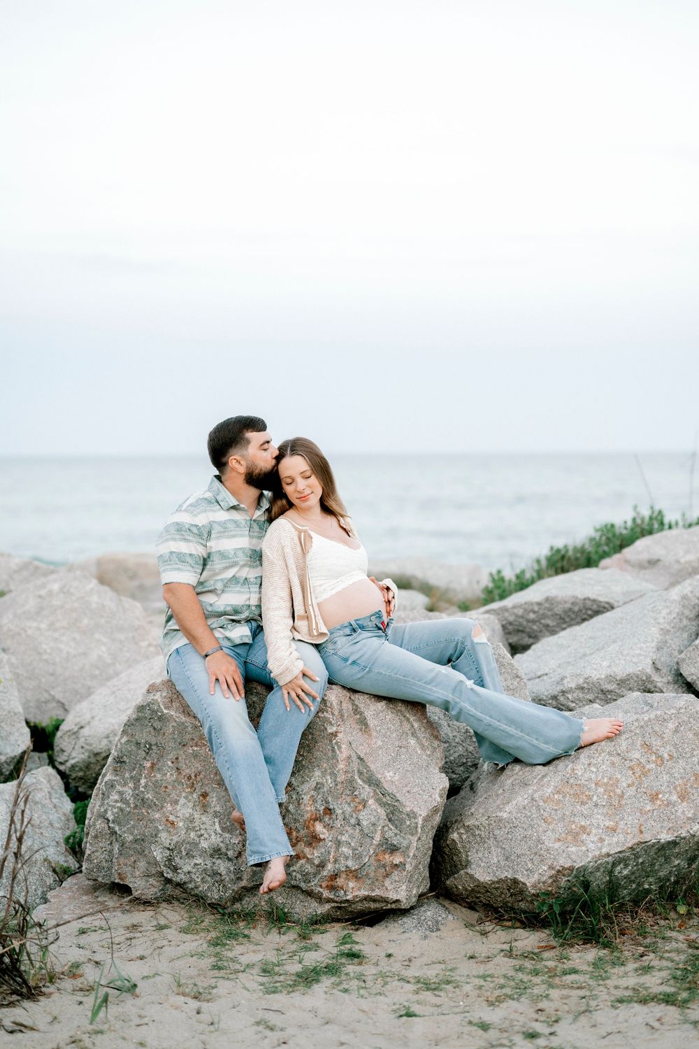 A couple sits closely on coastal rocks by the ocean, sharing a tender moment on a sandy beach.