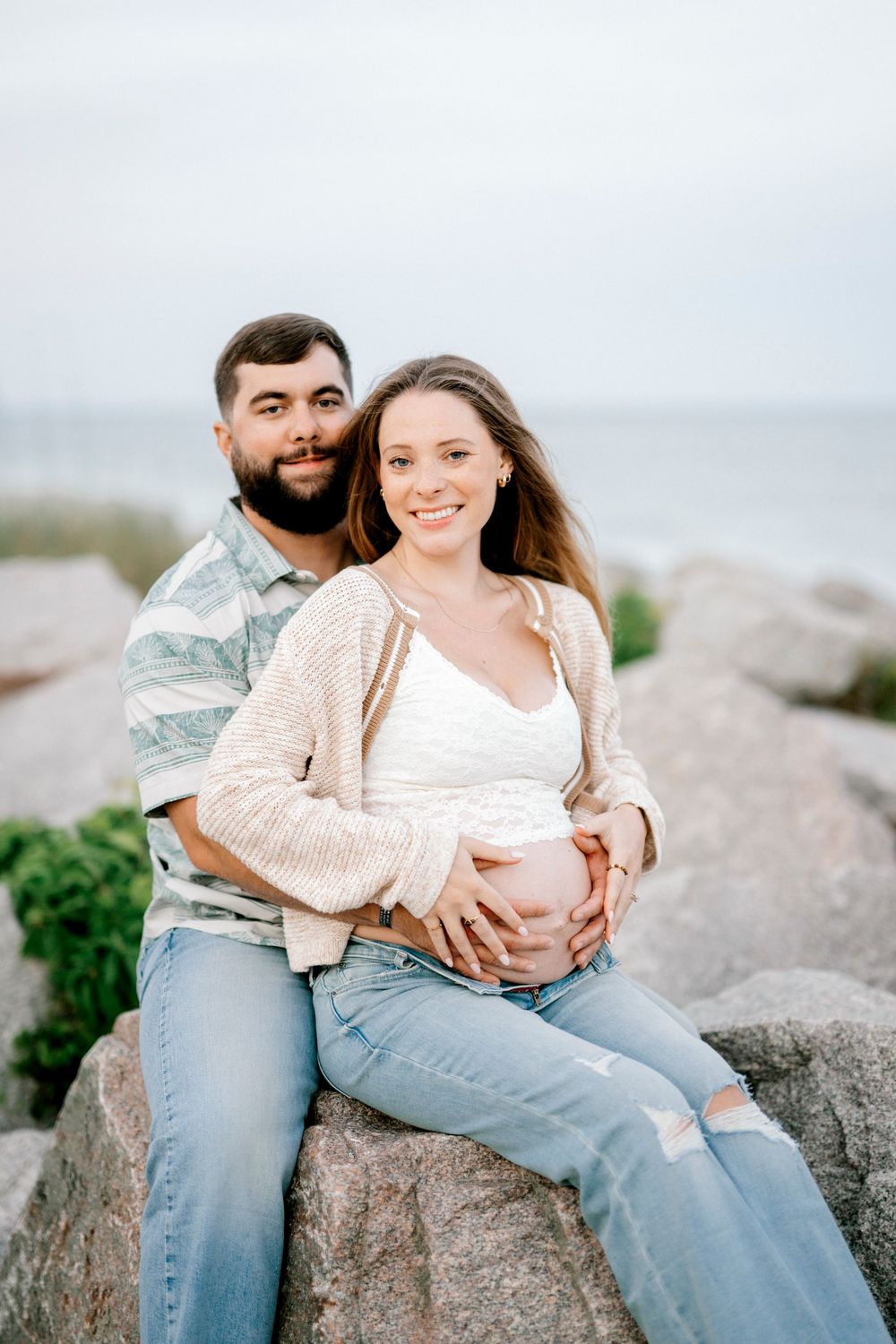 Couple sitting on rocks by the ocean, man embracing pregnant woman as they smile together outdoors.
