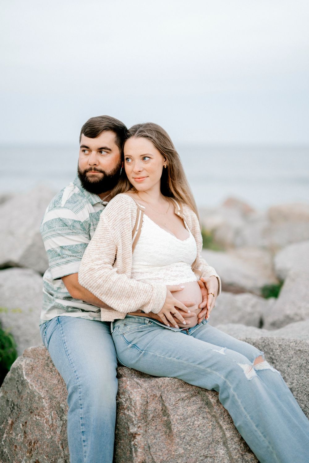 Pregnant couple sitting on rocks by the ocean, man embracing woman from behind as she holds her baby bump.