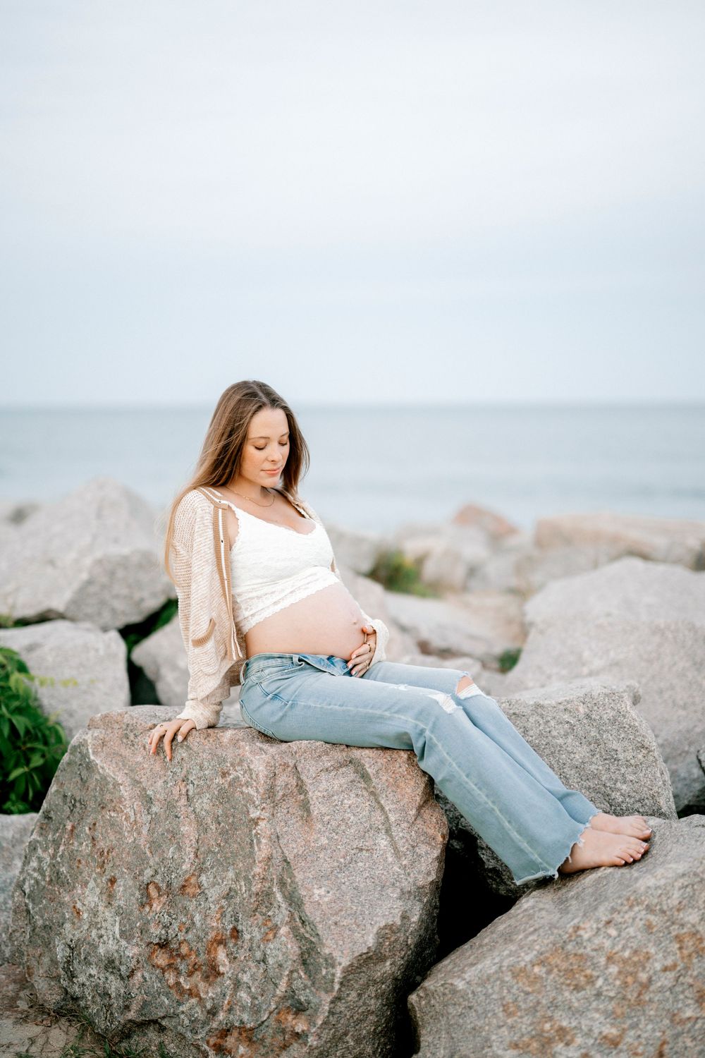 Pregnant woman sitting on rocks by the ocean, wearing jeans and white top, cradling her baby bump.