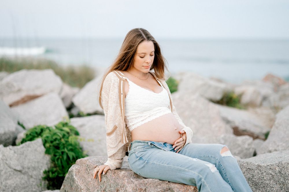 Pregnant woman sitting on rocks by the ocean, cradling her baby bump in a white top and jeans.