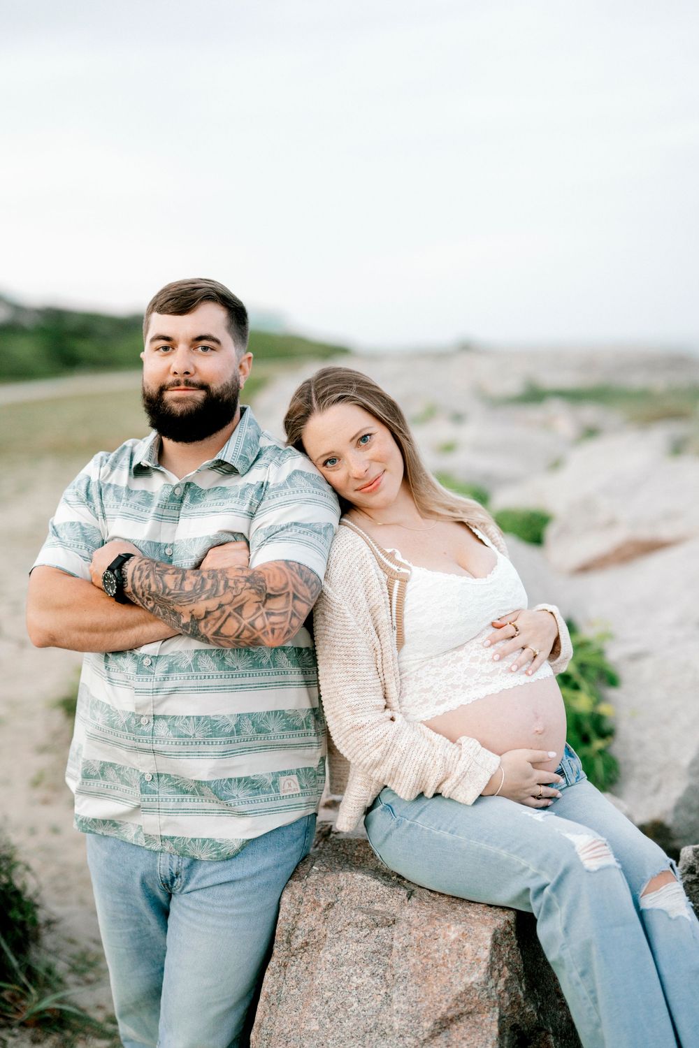 A bearded tattooed man and pregnant woman pose together on rocks outdoors in a maternity photo shoot.
