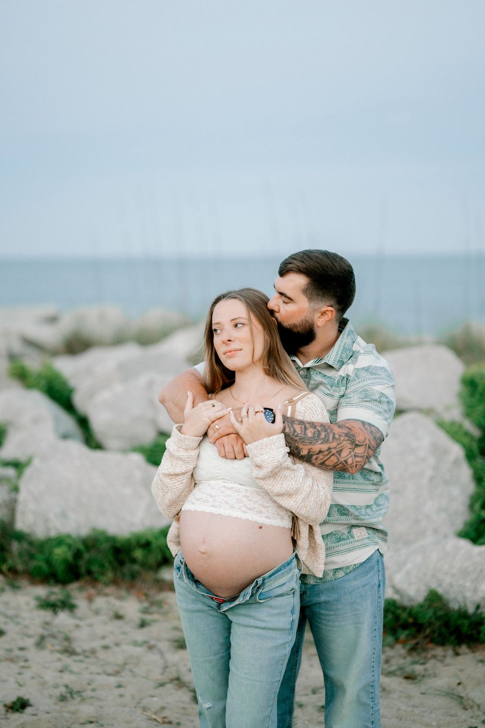 Pregnant couple embracing on beach, man with tattoos kissing woman's head while holding ultrasound photo near rocky shore.