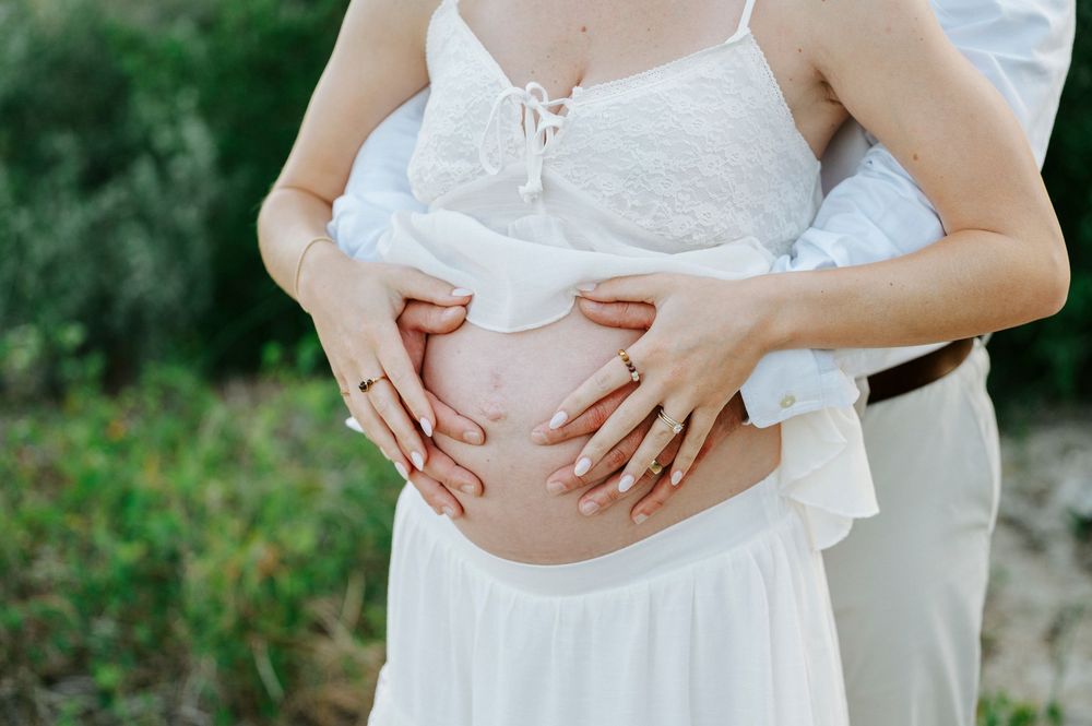 Pregnant woman in white dress with partner's hands cradling baby bump outdoors.