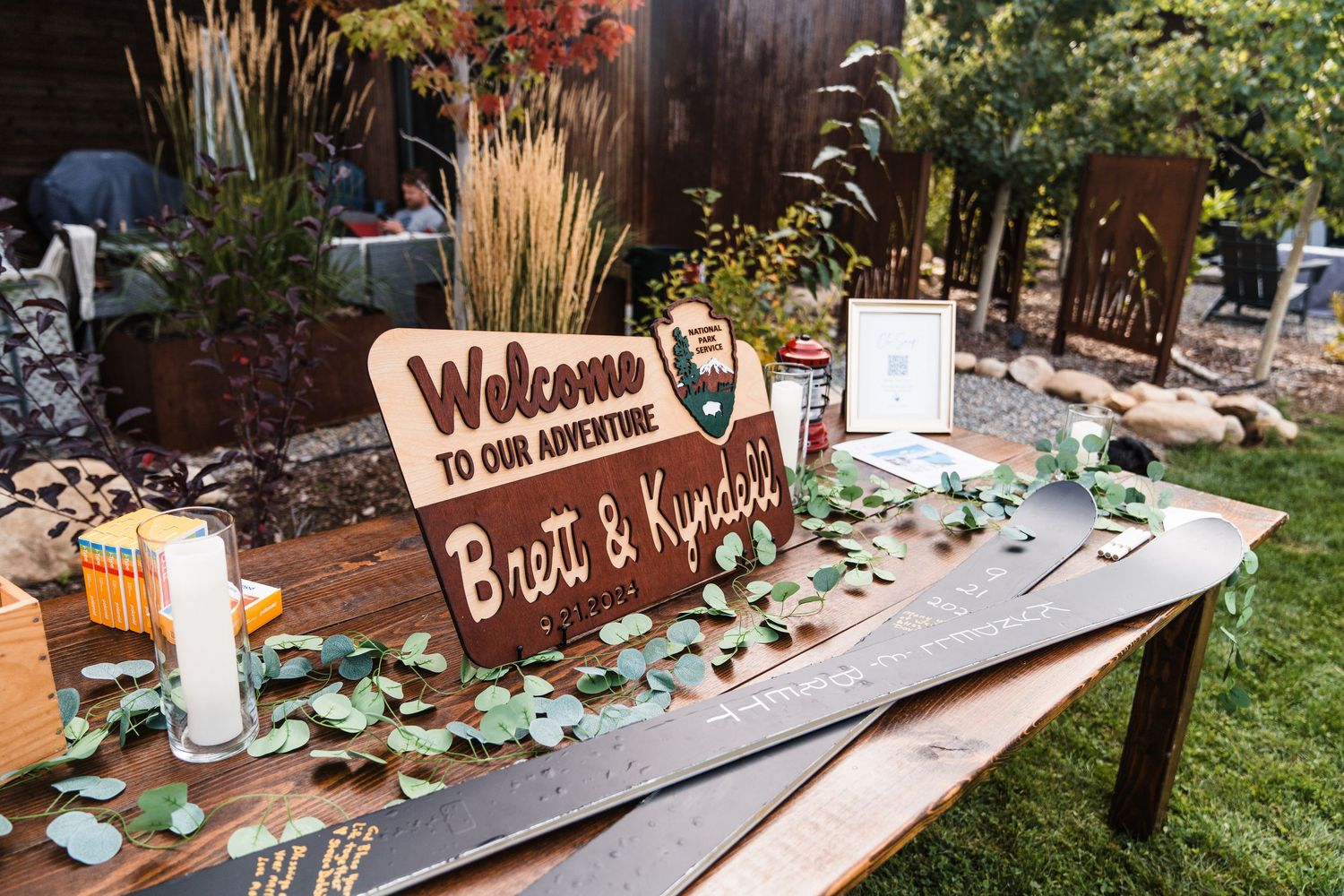 Rustic wedding welcome sign reading Brett & Kundis on a wooden table decorated with eucalyptus greenery and candles outdoors.