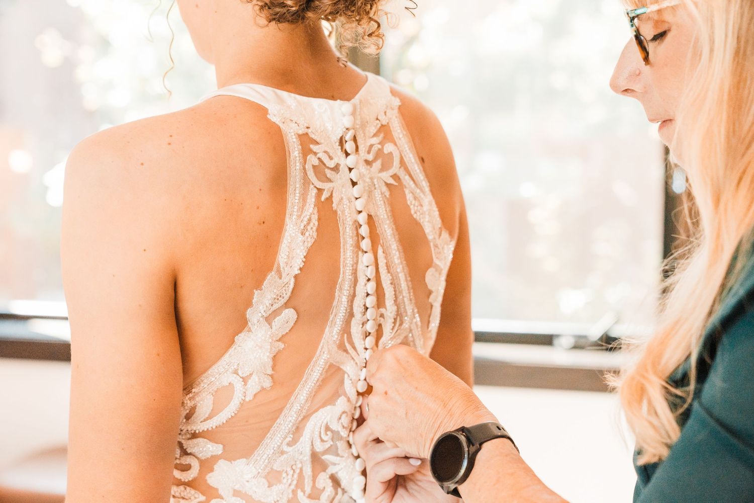 Bride in lace wedding dress getting buttoned up by a blonde woman in a bright indoor setting. north cascades elopement wedding