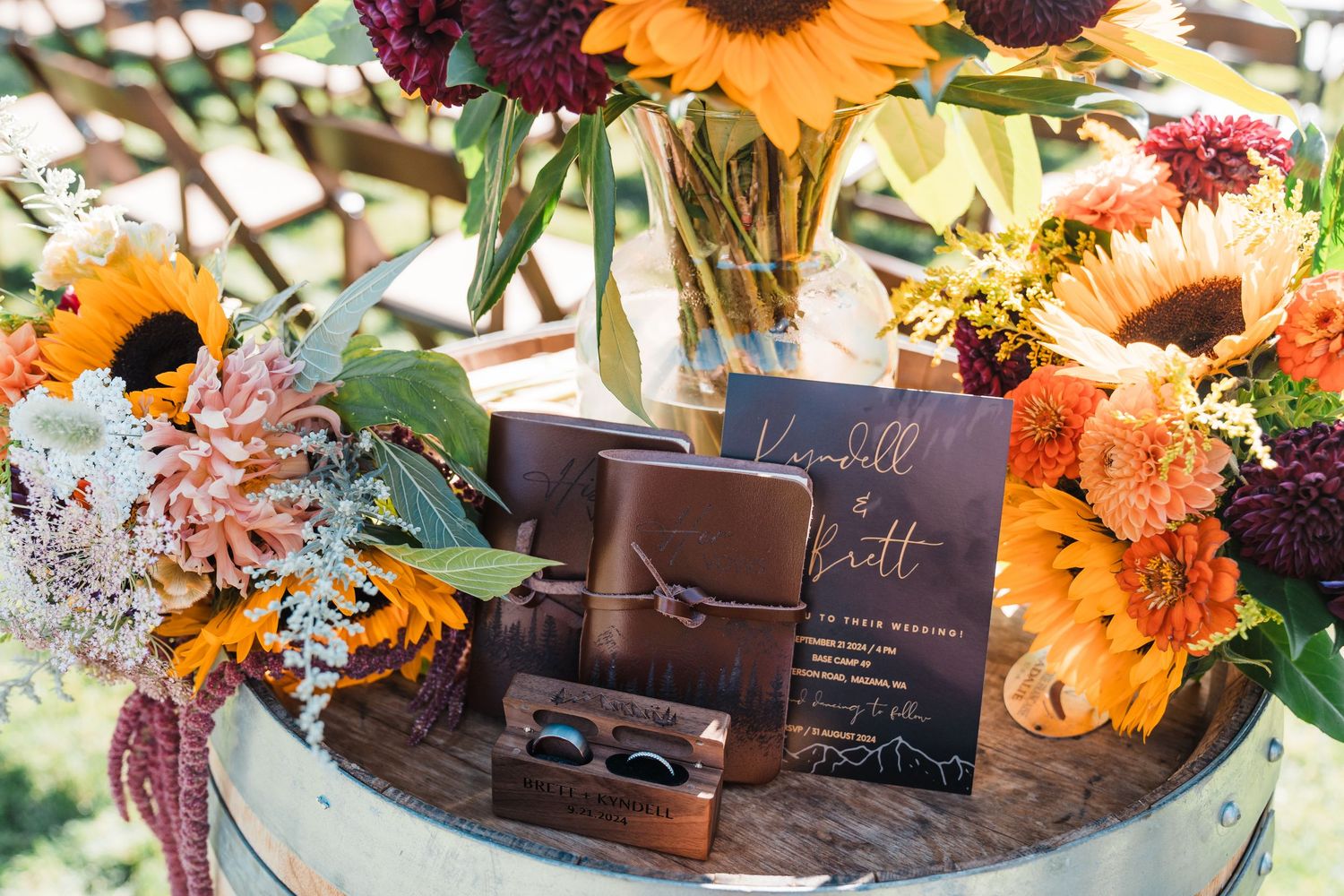 Rustic wedding display with sunflowers, dahlias, leather vow books, and wooden ring box on a barrel.