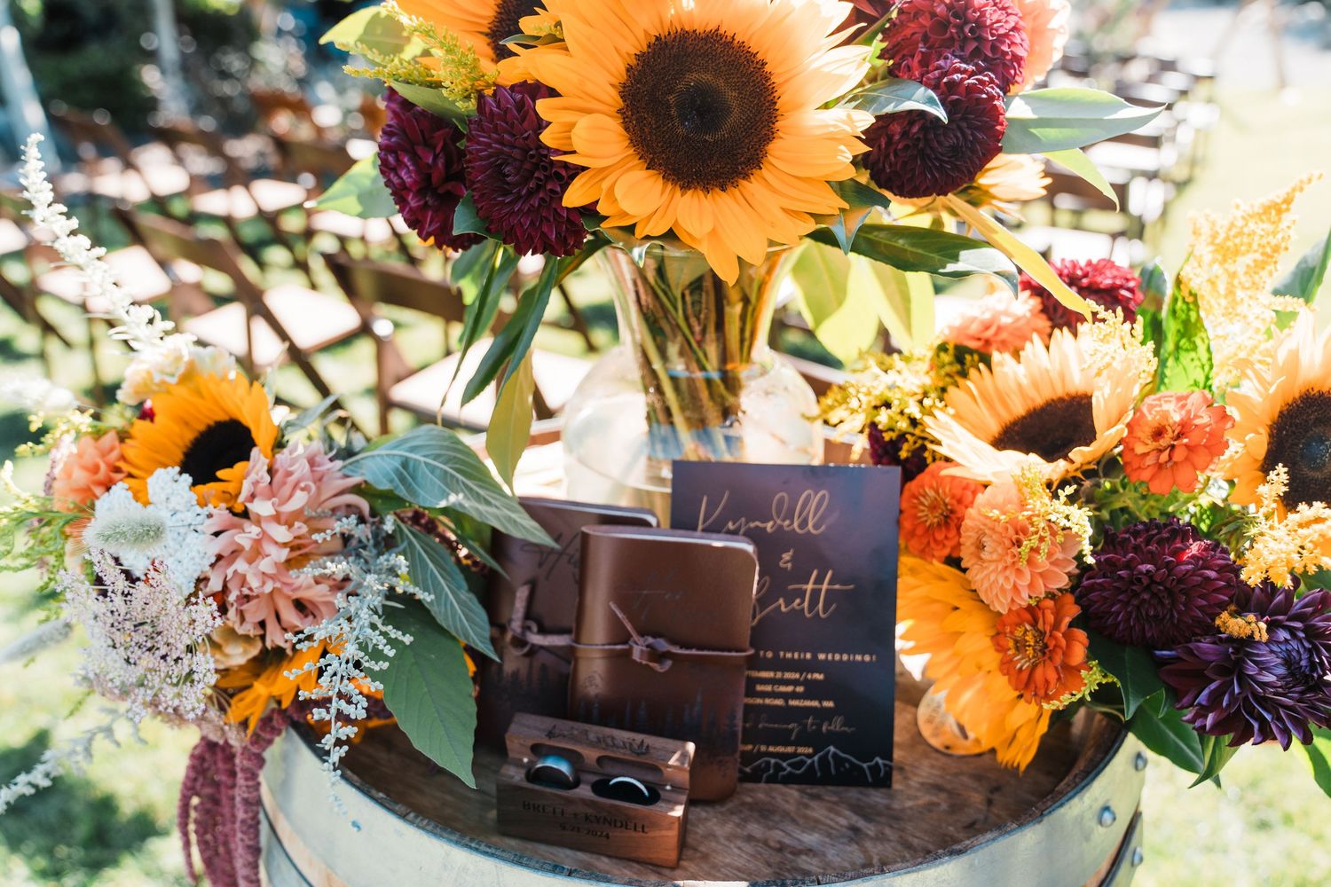 Fall wedding decor with sunflowers, burgundy dahlias, and orange blooms surrounding dark invitation suite on rustic barrel. north cascades e