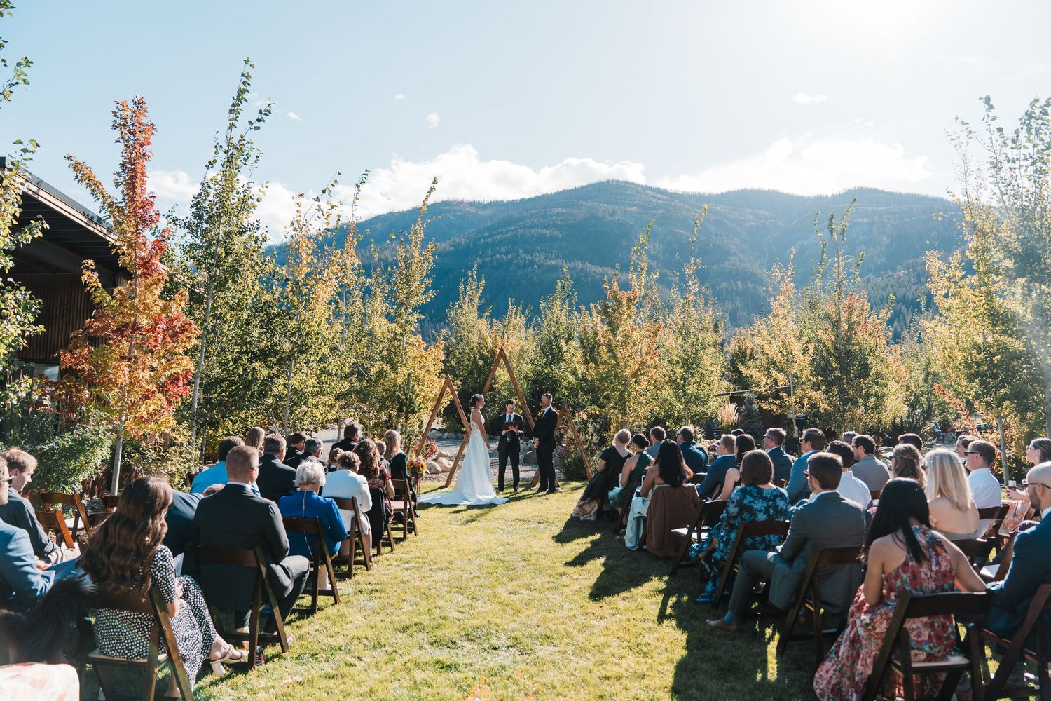 Outdoor wedding ceremony with guests seated on chairs facing couple at altar, surrounded by autumn trees and mountain backdrop. north cascad