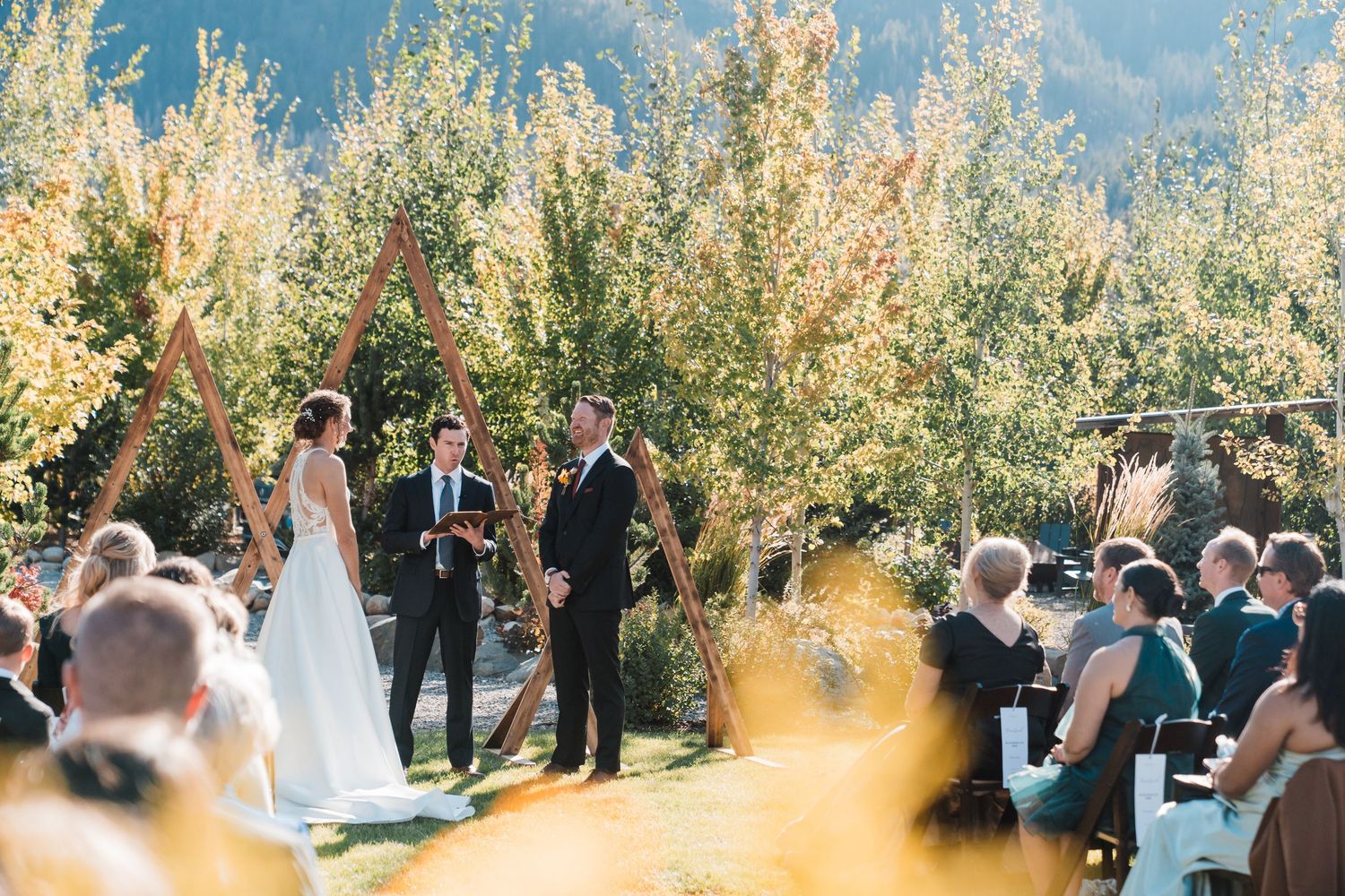 Outdoor wedding ceremony under a wooden triangular arch with mountain and autumn tree backdrop.north cascades elopement wedding