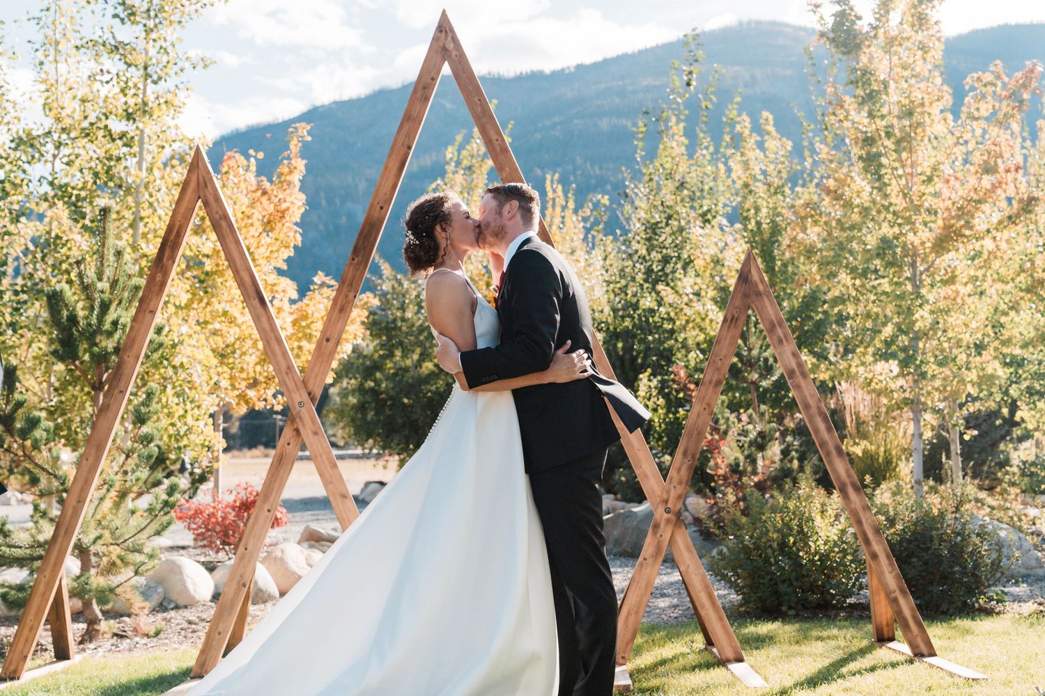 Bride and groom sharing a first kiss under wooden triangle arch with mountain backdrop. north cascades elopement wedding