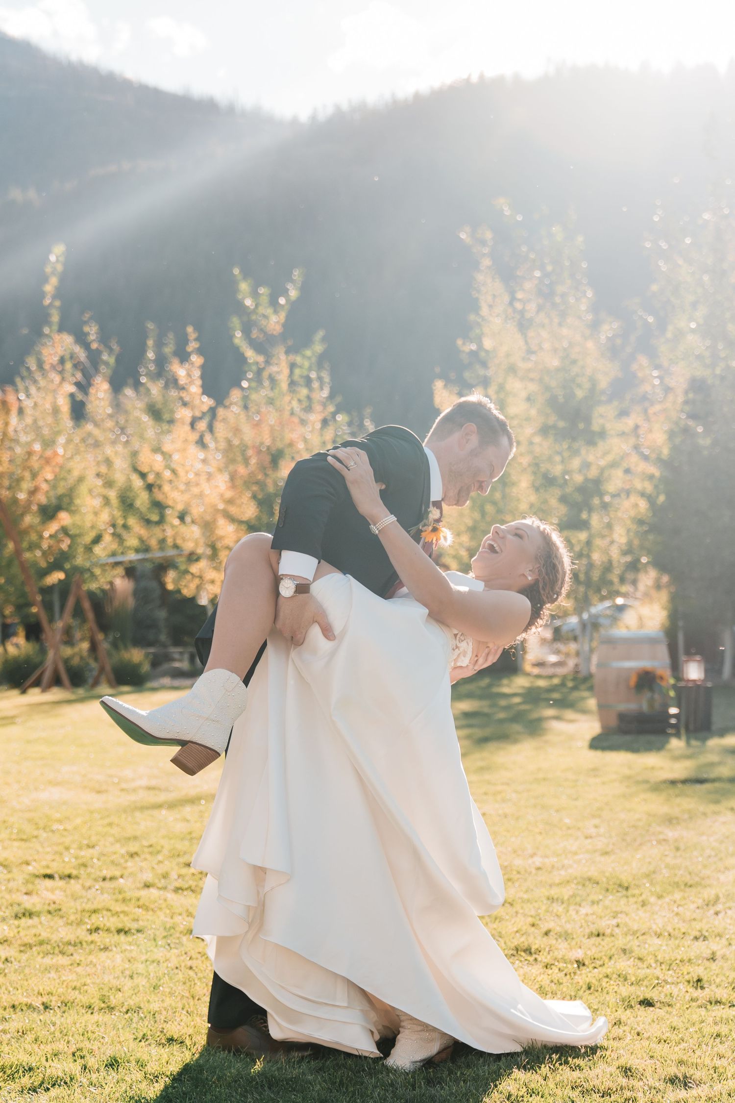 Groom lifting bride in white gown outdoors with sunlit mountain backdrop on their wedding day. north cascades elopement wedding