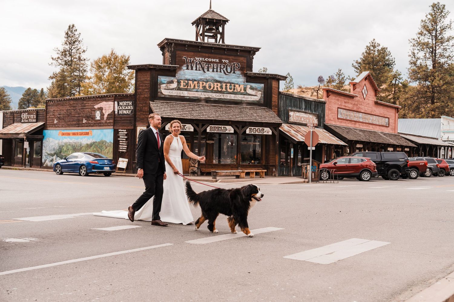 Bride and groom cross a street with their Bernese Mountain Dog in front of a rustic Western Emporium building. north cascades elopement wedd