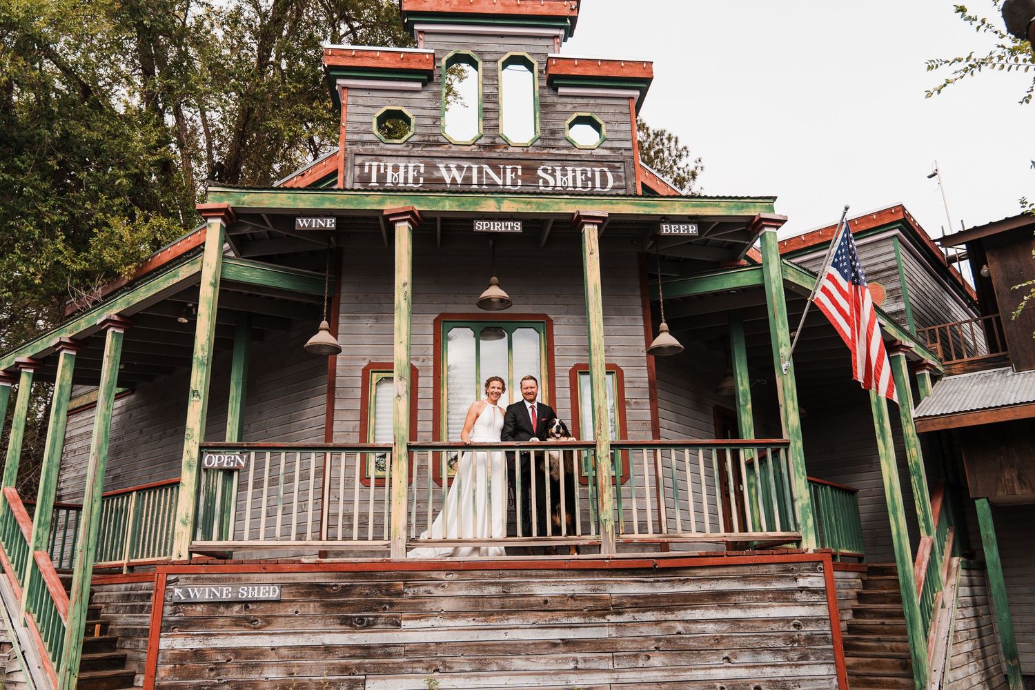 Couple poses on porch of rustic green Victorian Wine Shed building with American flag during wedding day. north cascades elopement wedding