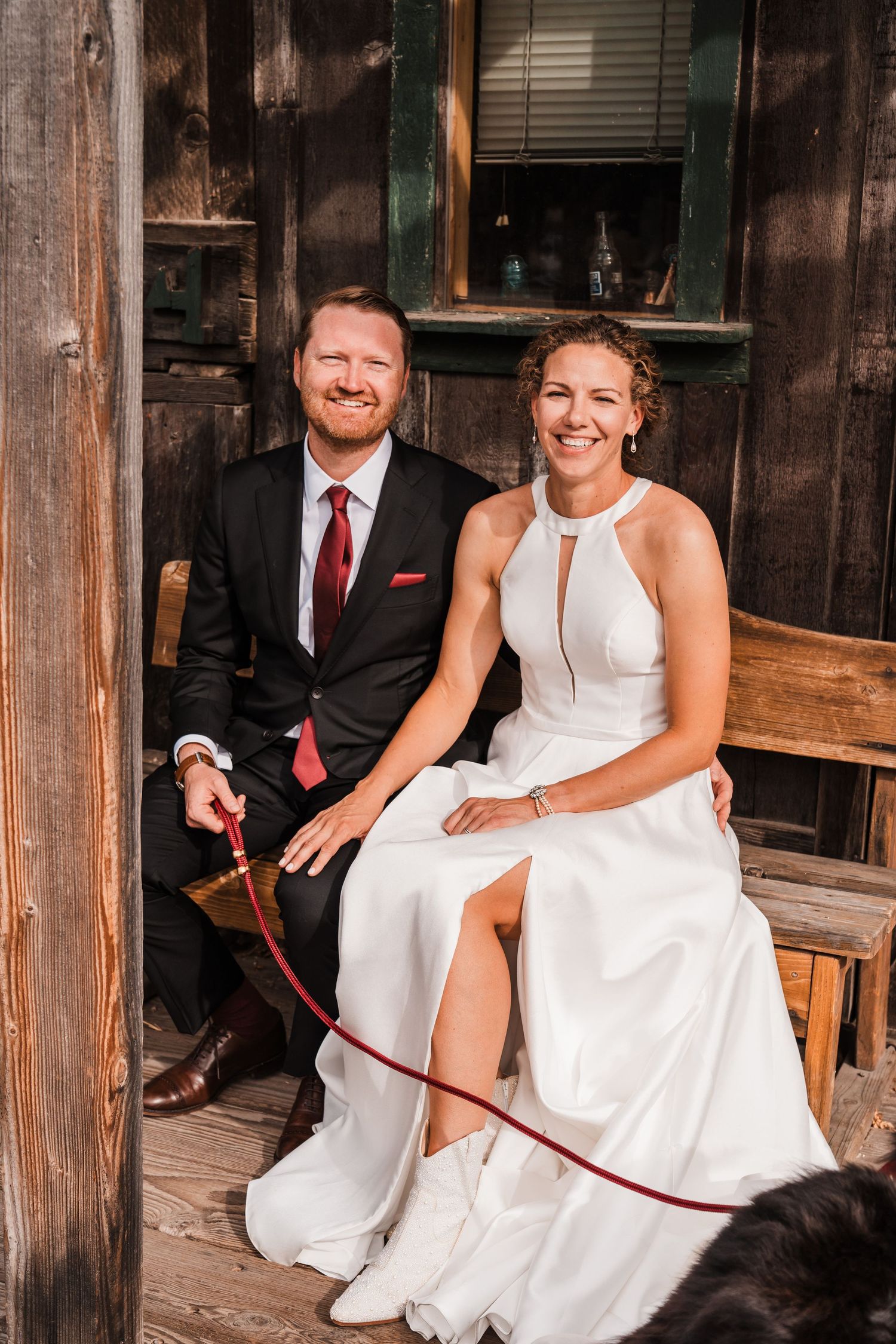 A smiling bride in a white halter gown and groom in a black suit with red tie sit together on a rustic wooden porch. north cascades elopemen