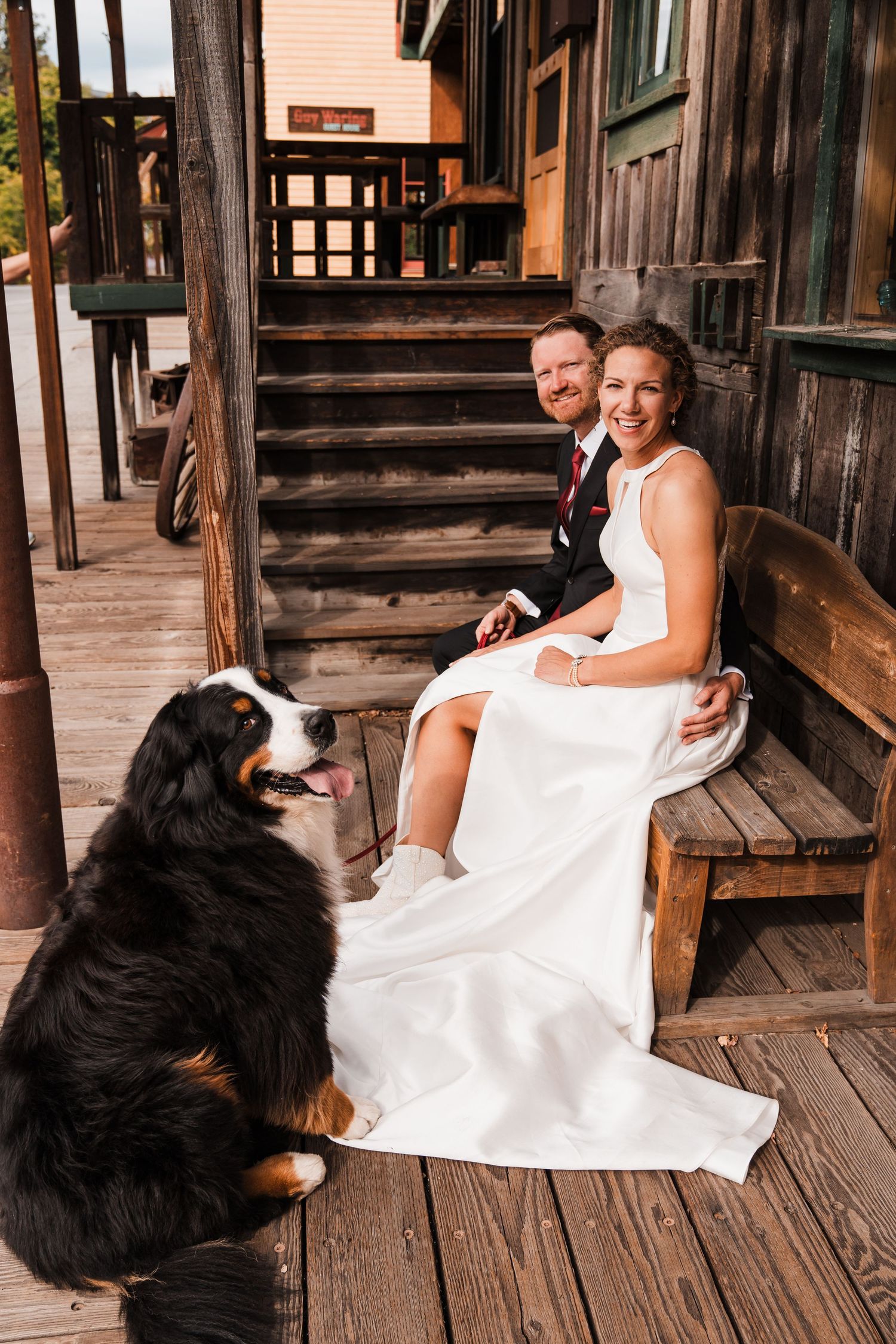 Bride and groom sit on wooden bench with Bernese Mountain Dog on rustic lodge porch in wedding attire. north cascades elopement wedding