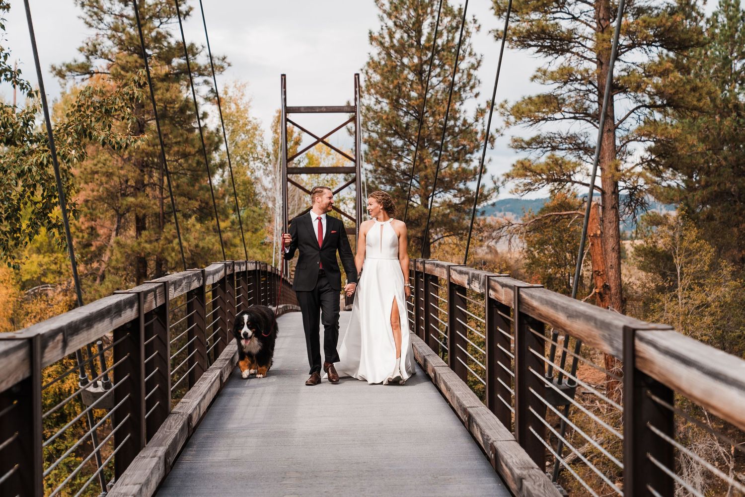 Bride and groom walking a dog on a wooden bridge surrounded by autumn trees at an outdoor wedding. north cascades elopement wedding