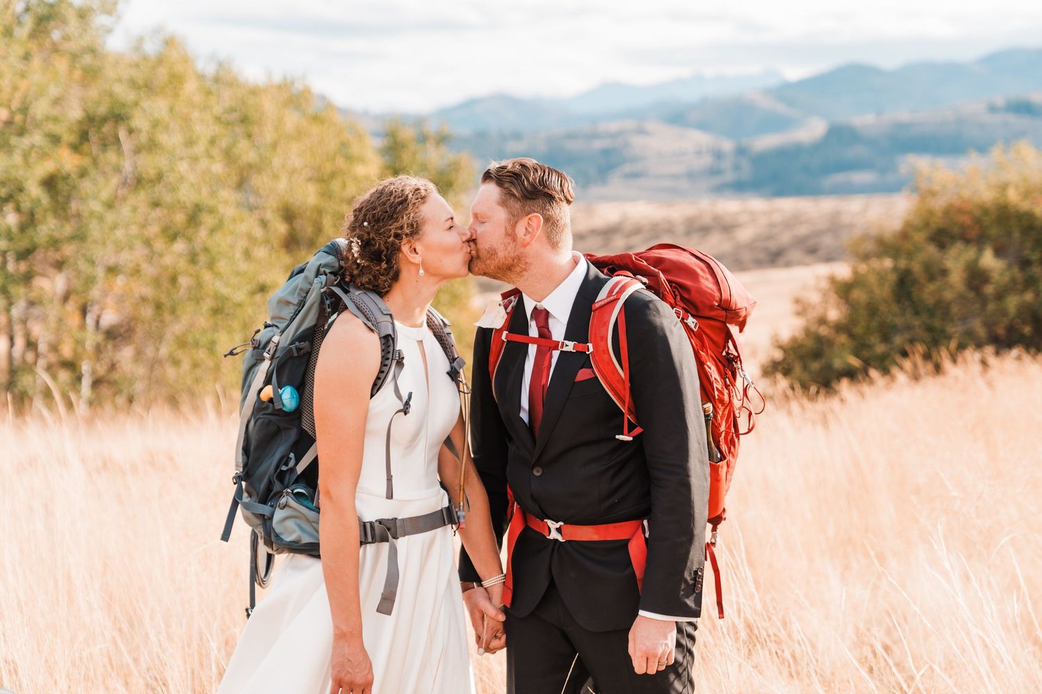 Bride and groom in wedding attire kissing in golden field while wearing red hiking backpacks.north cascades elopement wedding