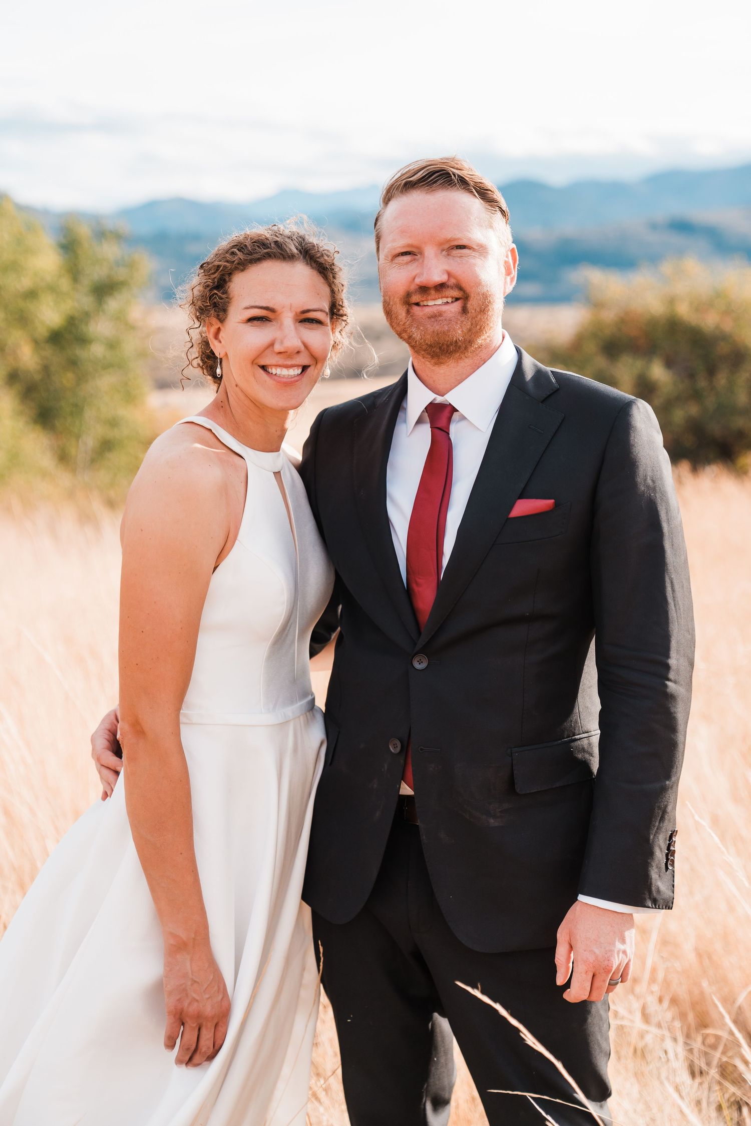 Smiling bride and groom pose together in golden field with mountain backdrop on wedding day. north cascades elopement wedding