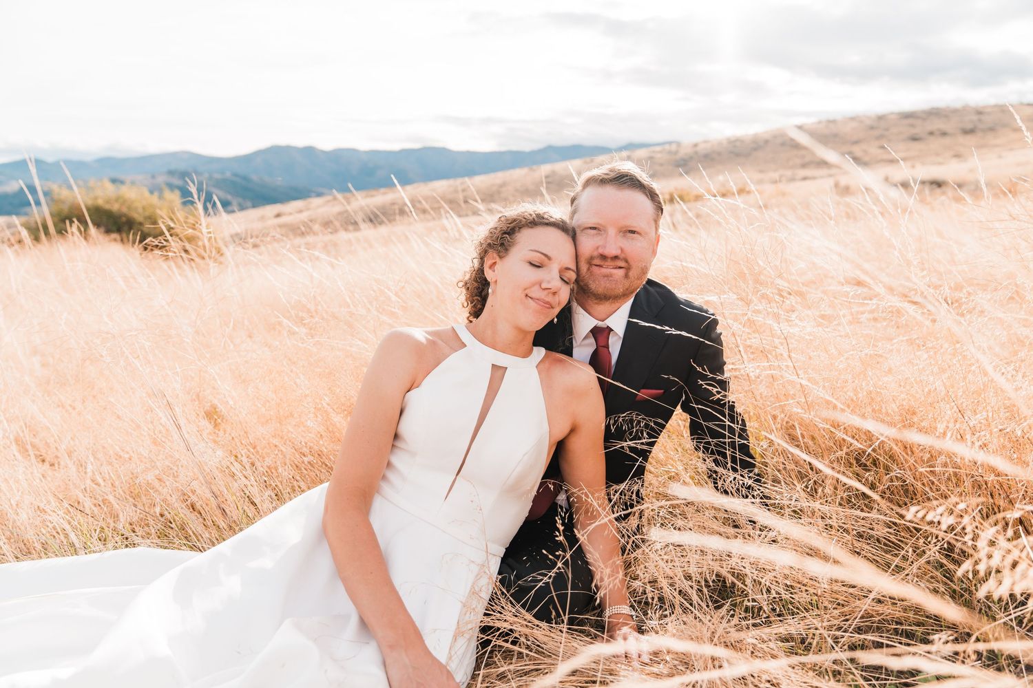 Bride and groom share tender moment sitting together in sunlit golden wheat field on wedding day. north cascades elopement wedding