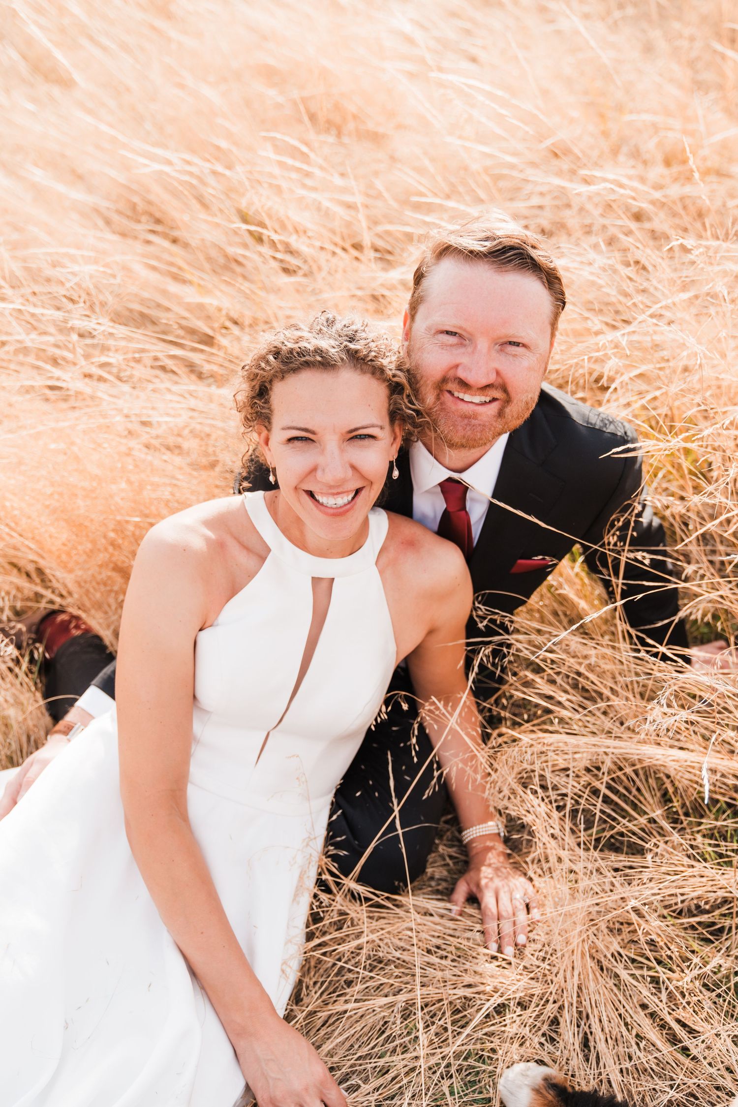 A smiling bride in a white halter dress and groom in a dark suit sit together in golden grass fields. north cascades elopement wedding