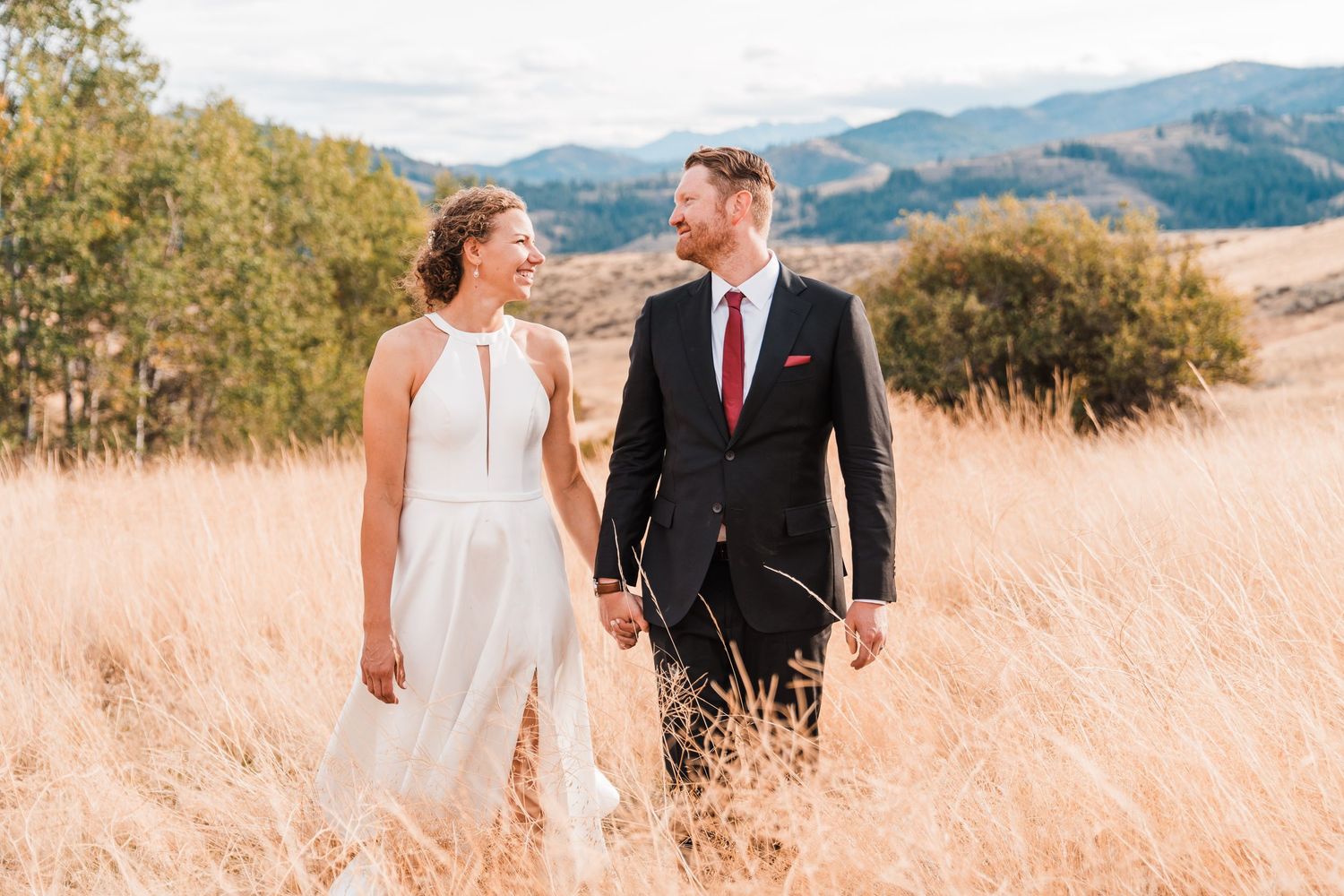 Bride and groom walk hand in hand through golden meadow with mountain backdrop.north cascades elopement wedding