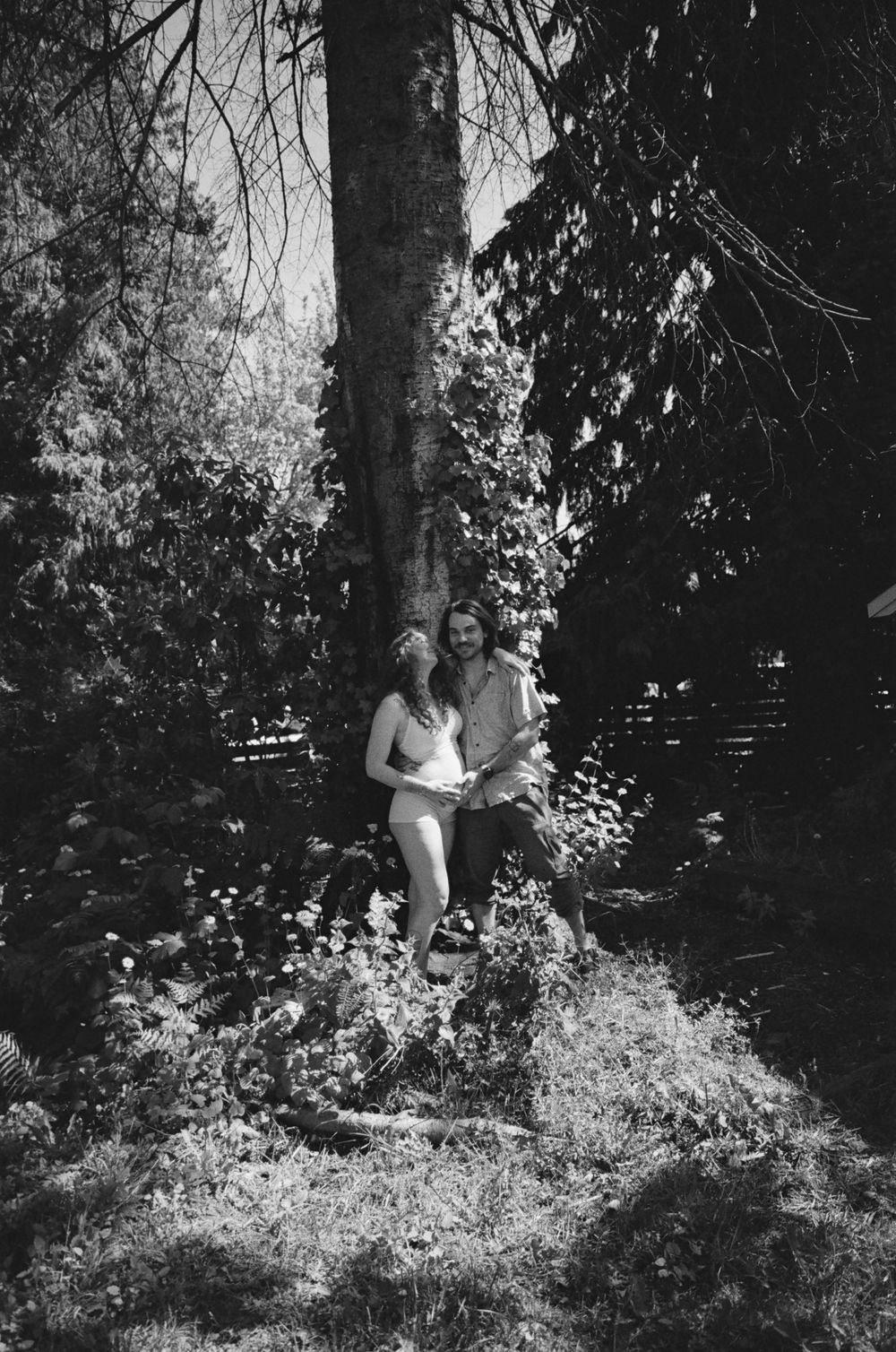 Black and white photo of a couple embracing near a large tree in a lush garden setting.
