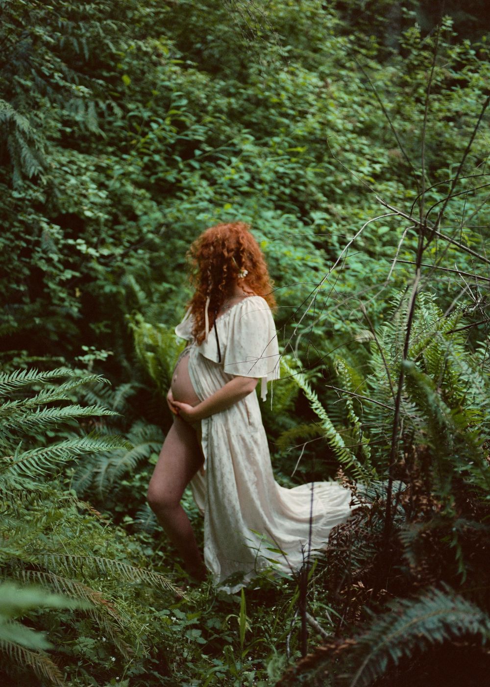 Pregnant redhead woman in sheer white dress cradles baby bump among lush green ferns in forest.