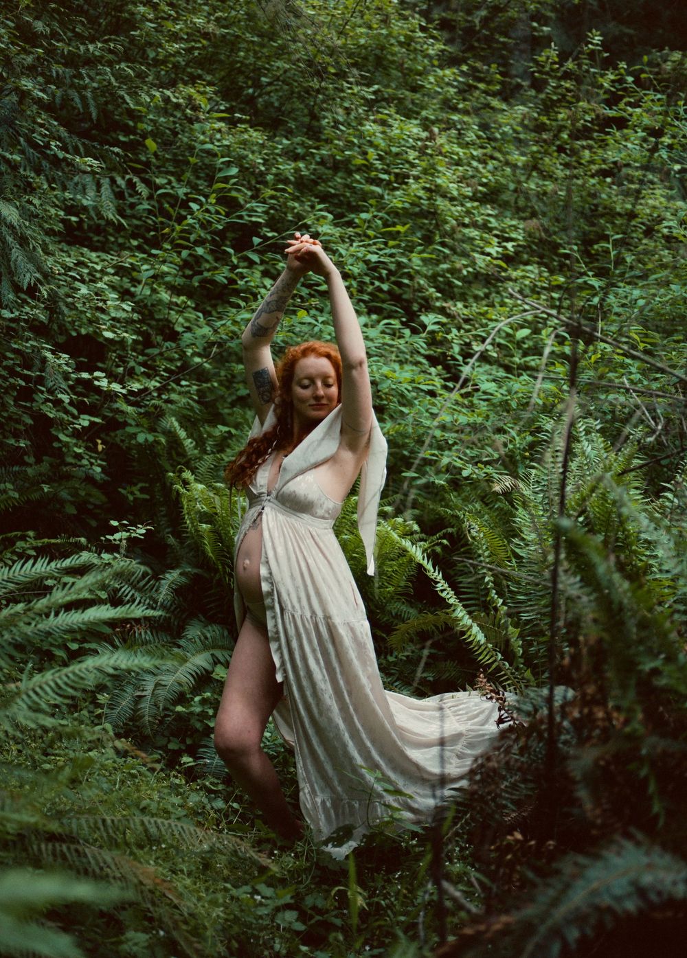Pregnant redhead woman in flowing white dress dancing among ferns in forest.